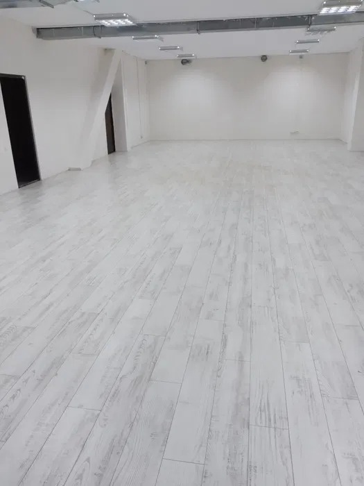 Jasne, bielone panele podłogowe w pustym, odnowionym biurze z białymi ścianami i surowym oświetleniem sufitowym.