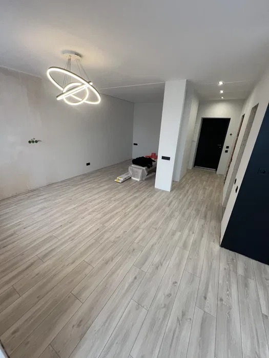 Wnętrze w trakcie prac wykończeniowych: położona podłoga imitująca drewno, surowe ściany z widocznymi puszkami elektrycznymi, nowoczesna lampa sufitowa i czarne drzwi w oddali.