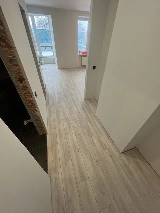 Jasne panele podłogowe ułożone wzdłuż korytarza, widok na okno i fragment ściany z surową cegłą po lewej stronie.