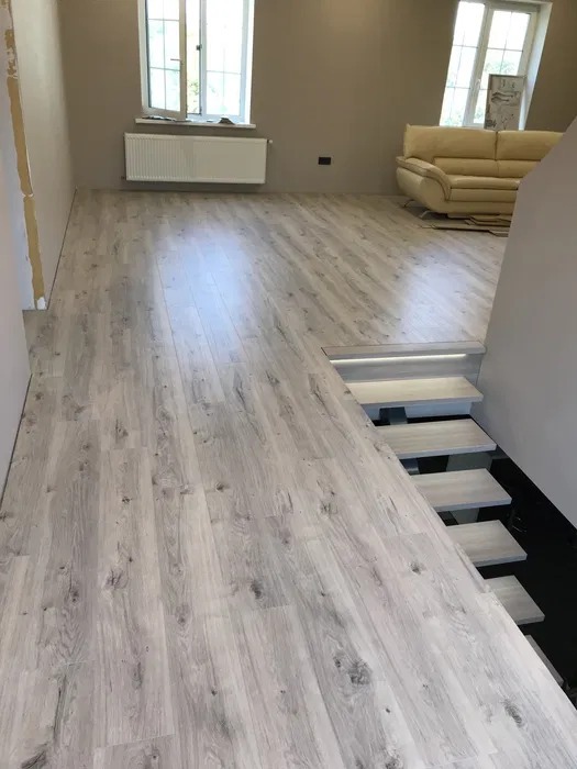 Jasnoszare panele podłogowe imitujące drewno, ułożone w nowym salonie z widokiem na schody z podświetleniem LED.