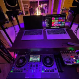 Stanowisko DJ-a z dwoma laptopami, mikserem Numark i oświetleniem scenicznym w fioletowej poświacie, w tle widoczni goście na przyjęciu.