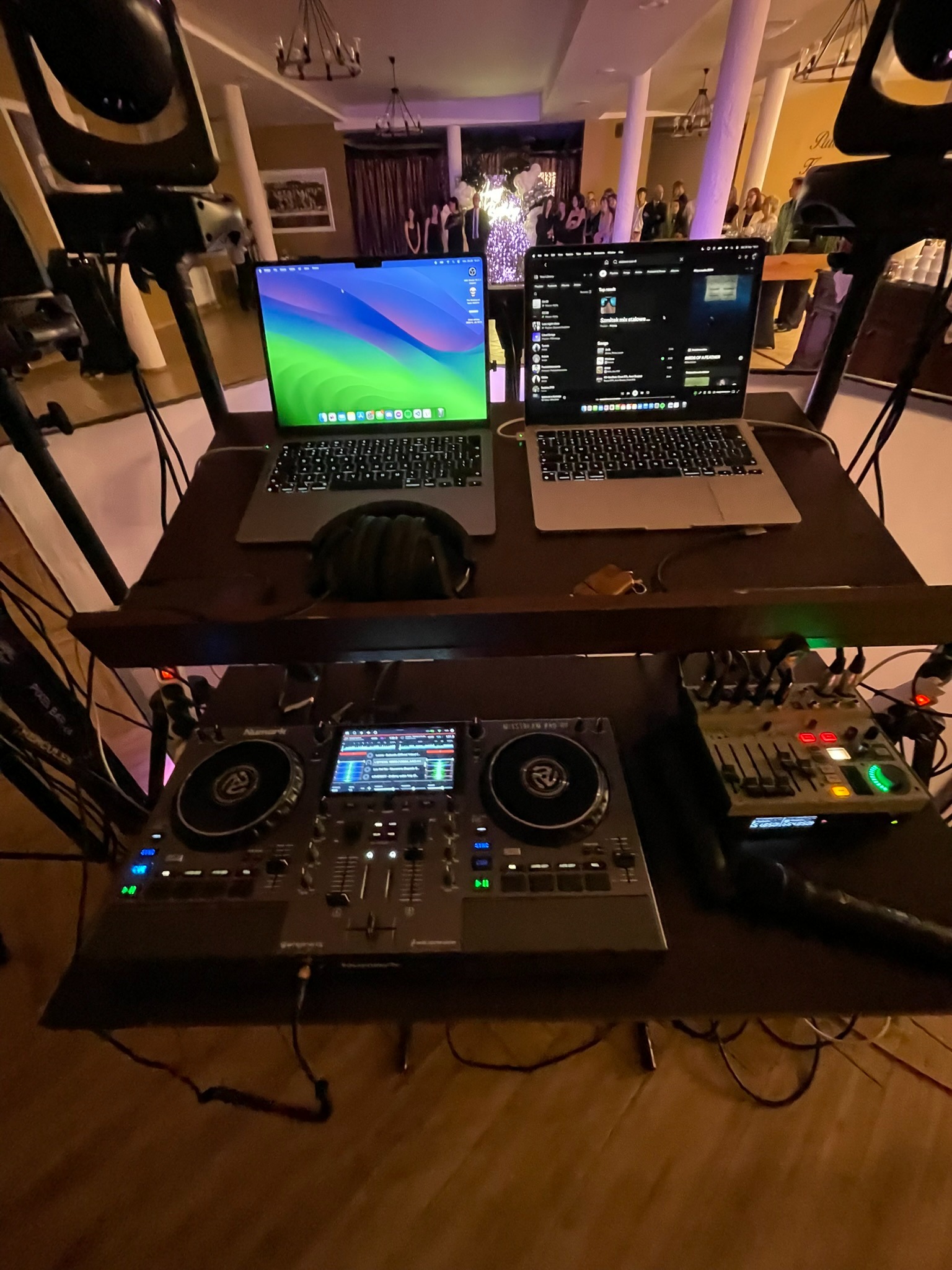 Stanowisko DJ-a z dwoma laptopami, słuchawkami, mikserem i kontrolerem DJ-skim na dwupoziomowym stole, w tle widoczni goście weselni.