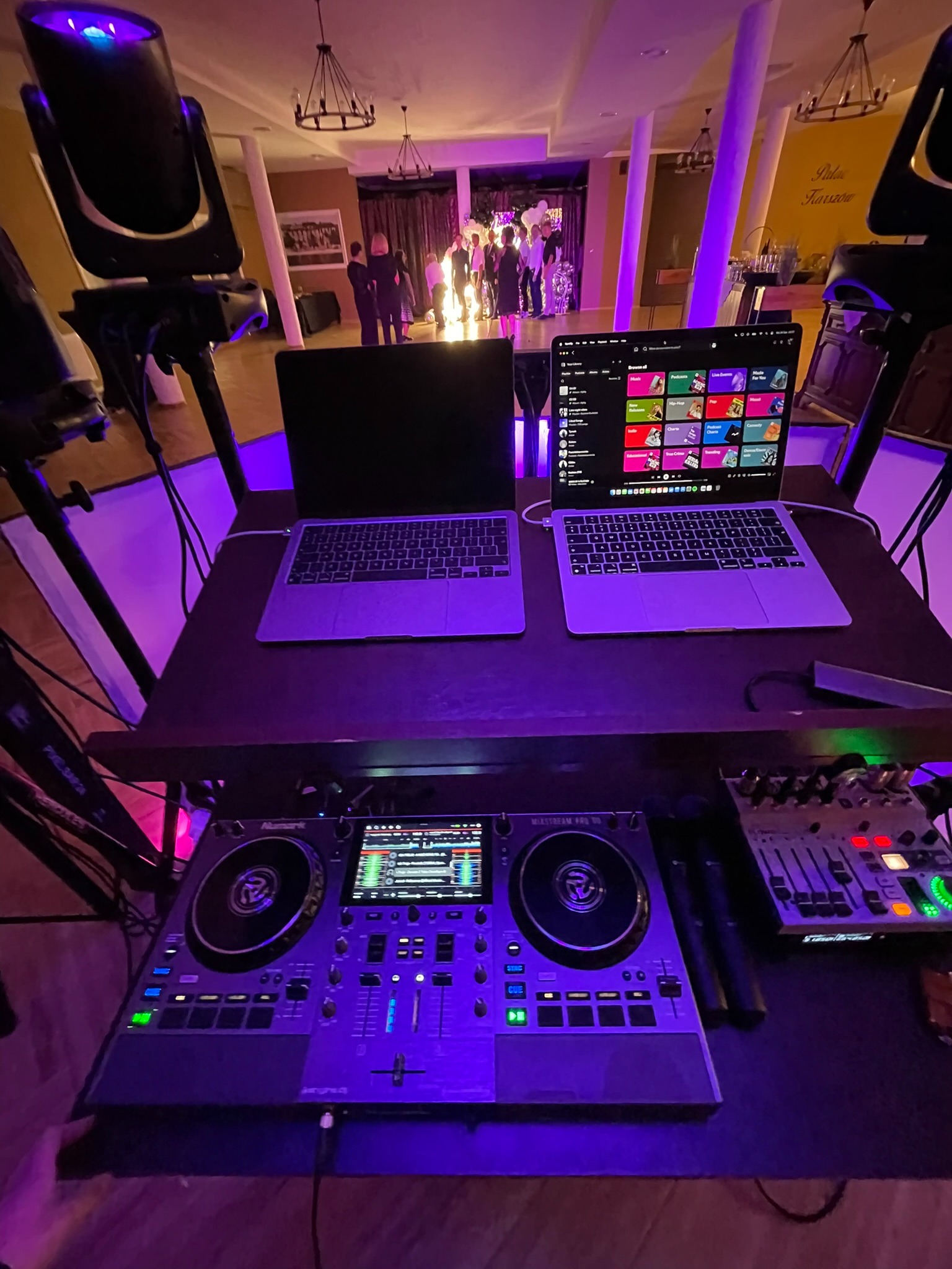Stanowisko DJ-a z dwoma laptopami, mikserem Numark i oświetleniem scenicznym w fioletowej poświacie, w tle widoczni goście na przyjęciu.