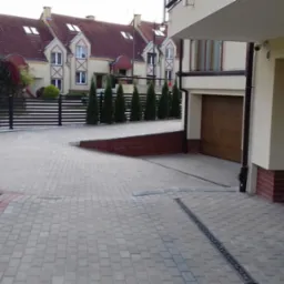 Nowoczesny podjazd z kostki brukowej w kolorze szarym, z widocznymi odpływami liniowymi, prowadzący do dwustanowiskowego garażu z drewnianą bramą, w tle ogrodzenie panelowe i rząd tuj.