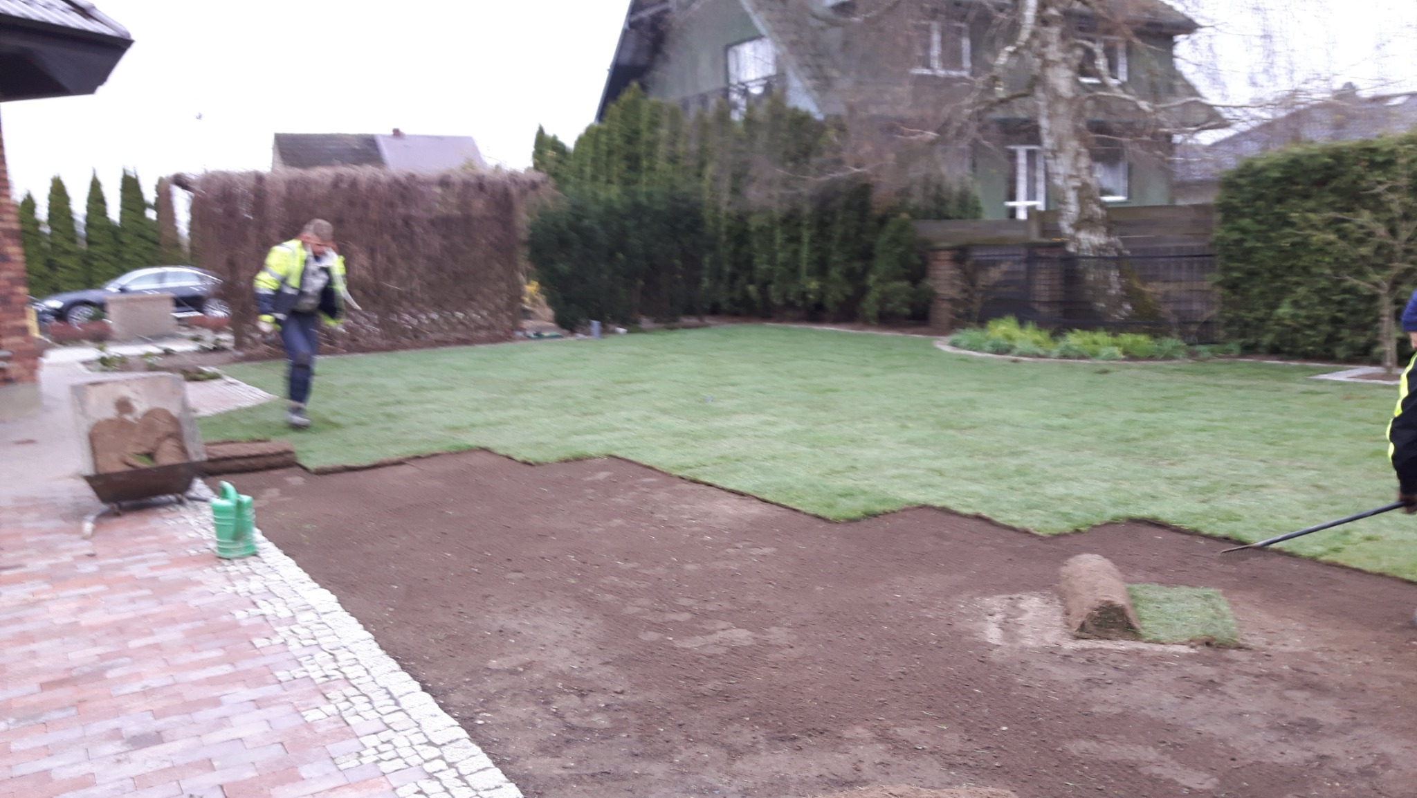 Rozkładanie trawy z rolki na przygotowanym podłożu, widoczny pracownik w odblaskowej kamizelce i narzędzia ogrodnicze, w tle dom z ogrodem.