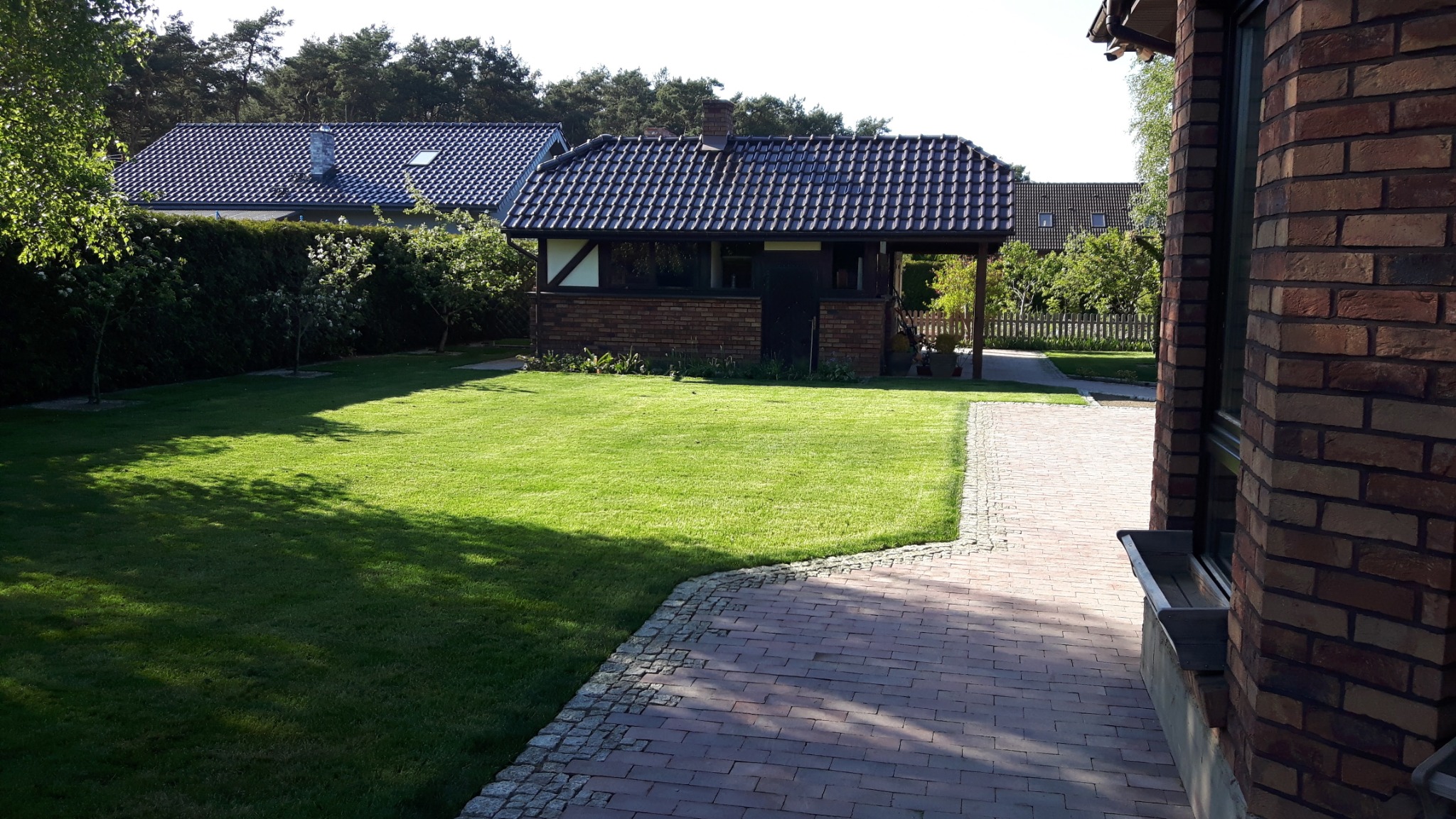 Zadbany trawnik z widokiem na ceglany budynek gospodarczy z ciemną dachówką, obok ścieżka z kostki brukowej i fragment ściany budynku mieszkalnego z cegły.