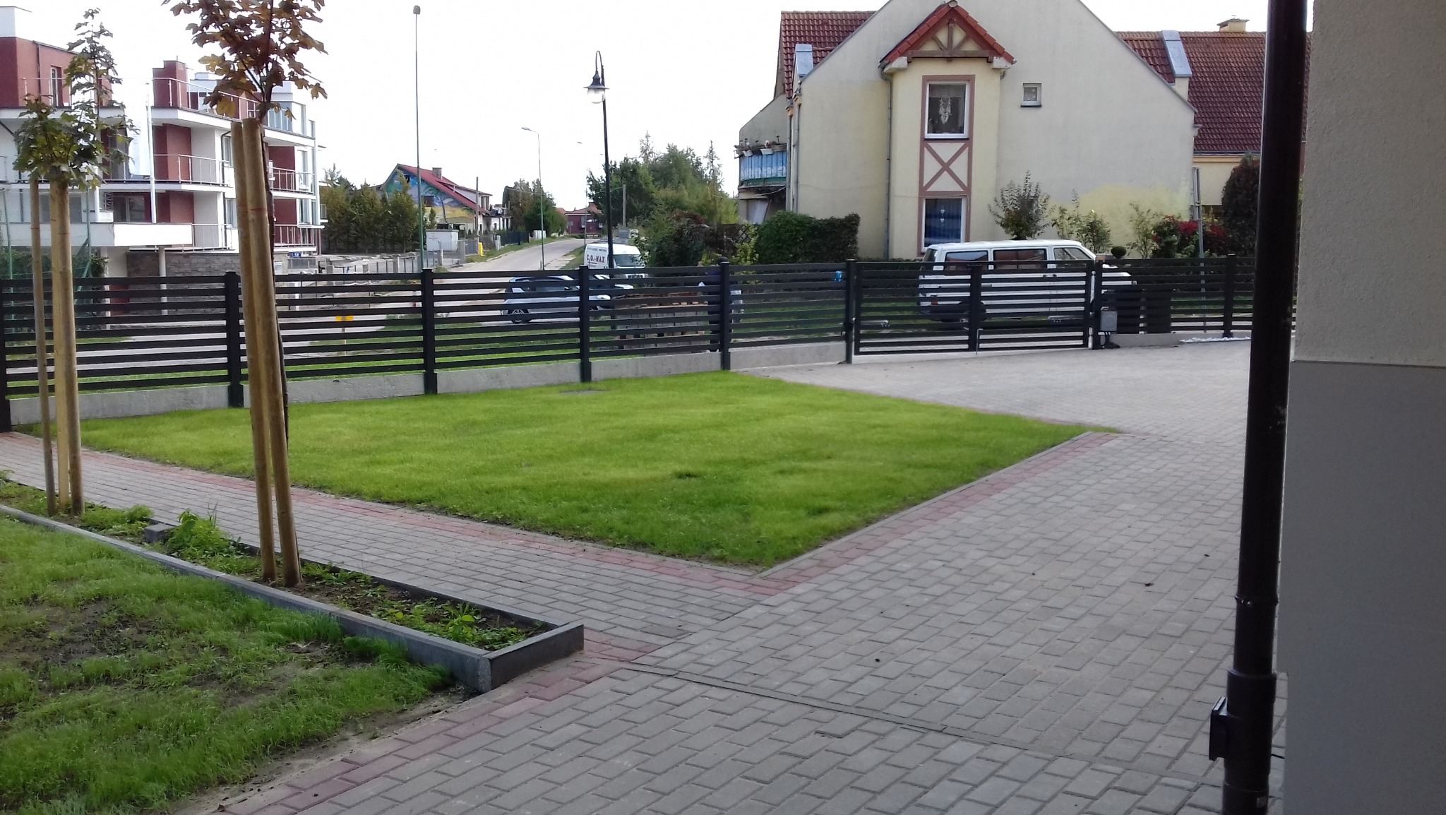 Nowoczesne, poziome ogrodzenie panelowe w kolorze antracytowym, otaczające równo przystrzyżony trawnik, z kostką brukową i młodymi drzewkami w tle zabudowy mieszkaniowej.