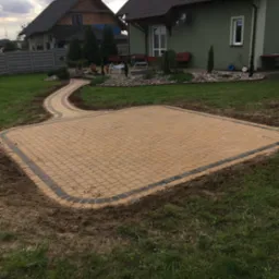 Żółta kostka brukowa ułożona w kwadratowy wzór z ciemniejszą obwódką, połączona ścieżką z kostki do domu z zieloną elewacją i zadbanym ogrodem.