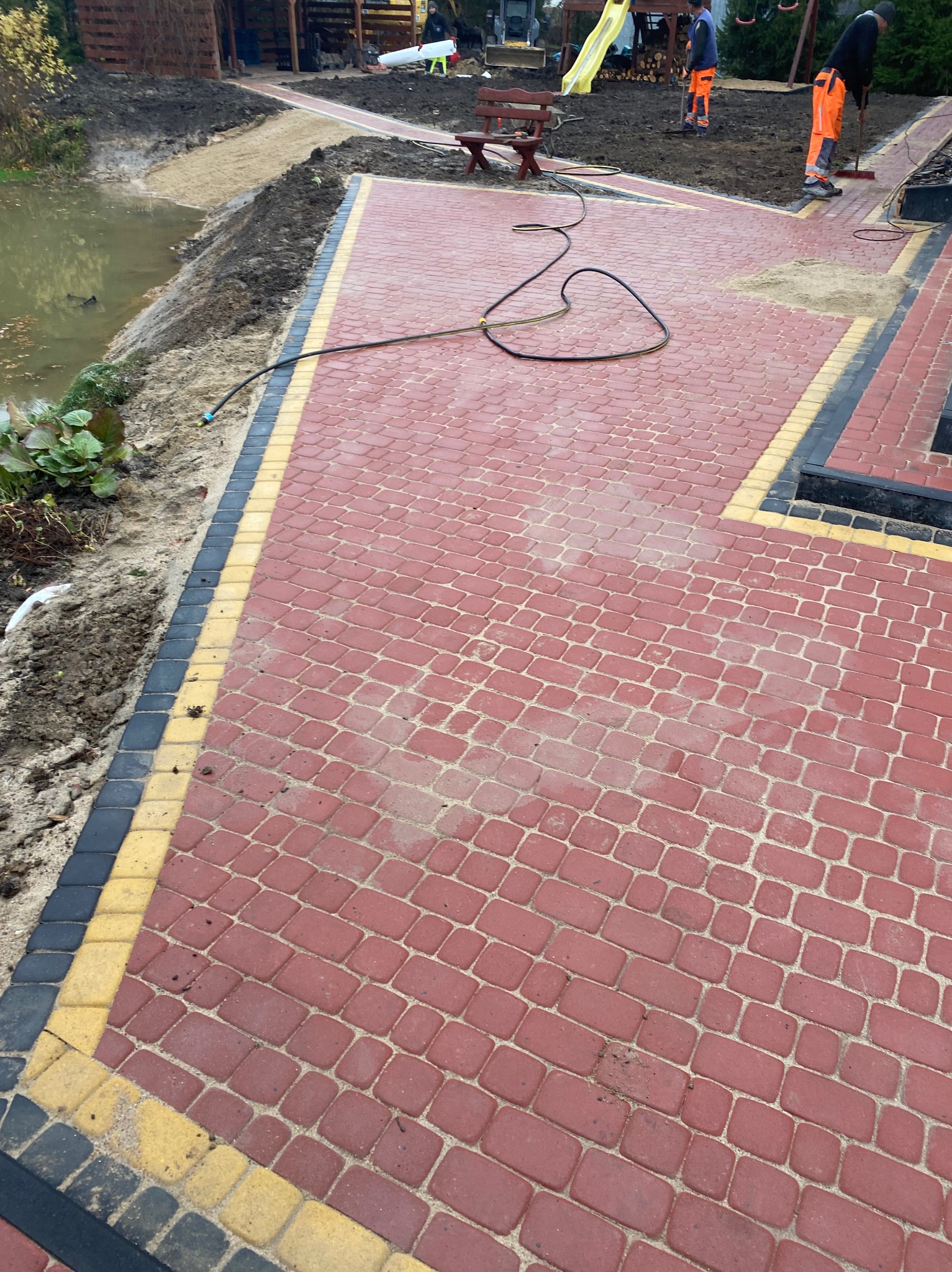 Czerwona kostka brukowa z żółto-czarnym obrzeżem, świeżo ułożona, obok staw i dwóch pracowników w pomarańczowych kombinezonach zamiatających piasek w tle.