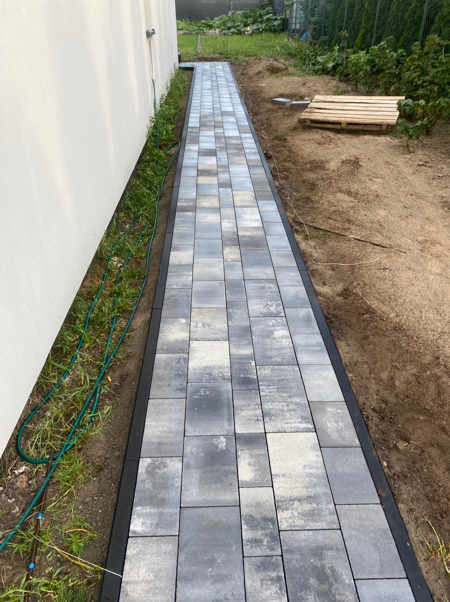 Ścieżka z szarej kostki brukowej prowadząca wzdłuż białej ściany budynku, obrzeża z ciemnego kamienia, widoczny trawnik i drewniana paleta w tle.