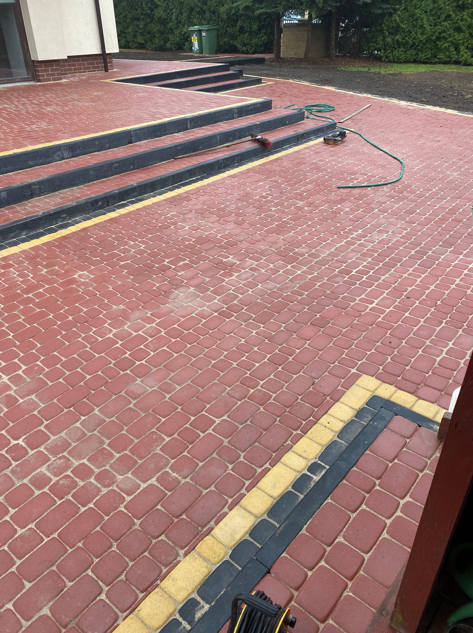 Czerwona kostka brukowa ułożona na podjeździe z czarnymi obrzeżami i żółtymi akcentami, prowadząca do schodów wejściowych. W tle widoczna zieleń.