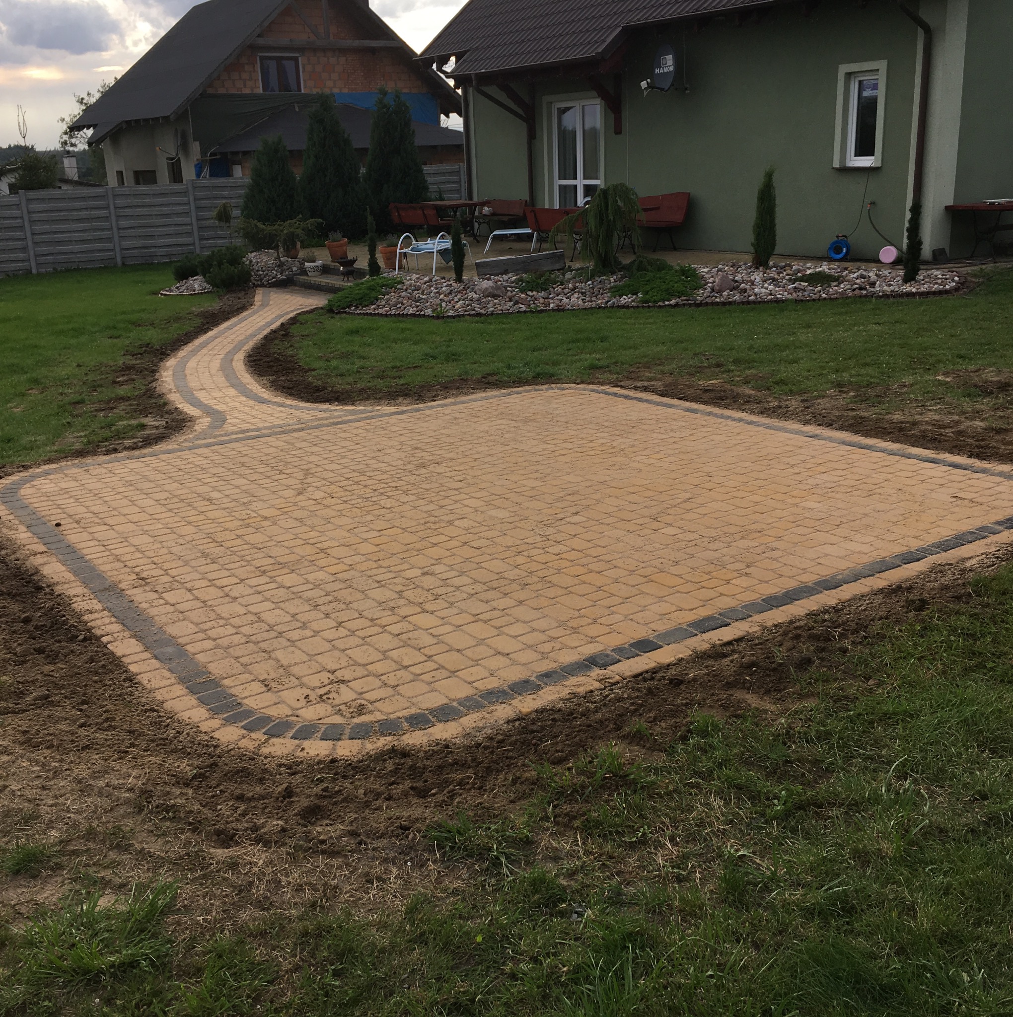 Żółta kostka brukowa ułożona w kwadratowy wzór z ciemniejszą obwódką, połączona ścieżką z kostki do domu z zieloną elewacją i zadbanym ogrodem.