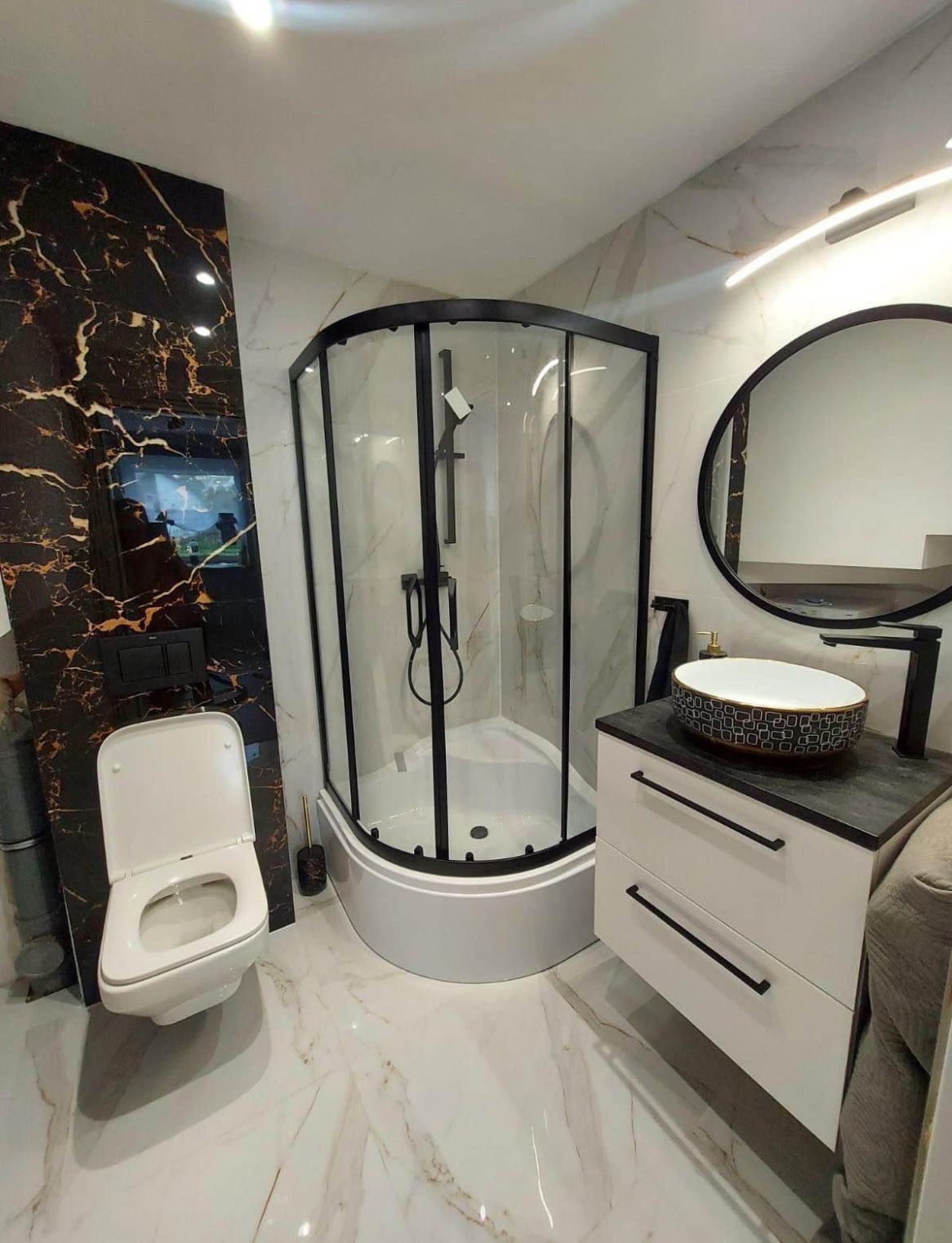 Nowoczesna łazienka z kabiną prysznicową z czarnymi profilami, wiszącą toaletą, umywalką nablatową w geometryczne wzory i marmurową okładziną ścian w odcieniach bieli i czerni ze złotymi żyłami.