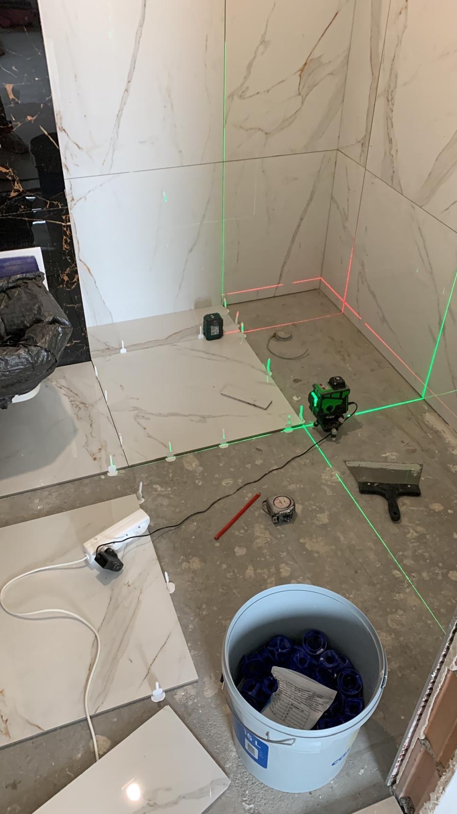 Montaż płytek w łazience z użyciem poziomicy laserowej, widoczne płytki imitujące marmur, wiadro z krzyżykami dystansowymi, szpachelka i ołówek.