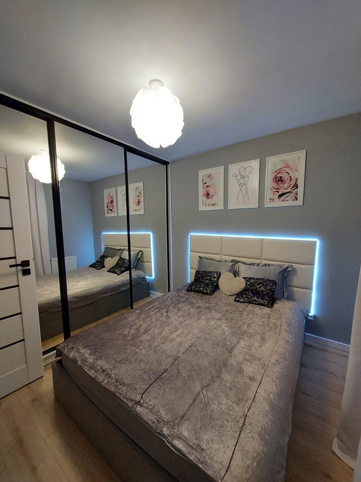 Sypialnia z łóżkiem z podświetlanym zagłówkiem, lustrzaną szafą przesuwną i dekoracyjnymi obrazkami na szarej ścianie.