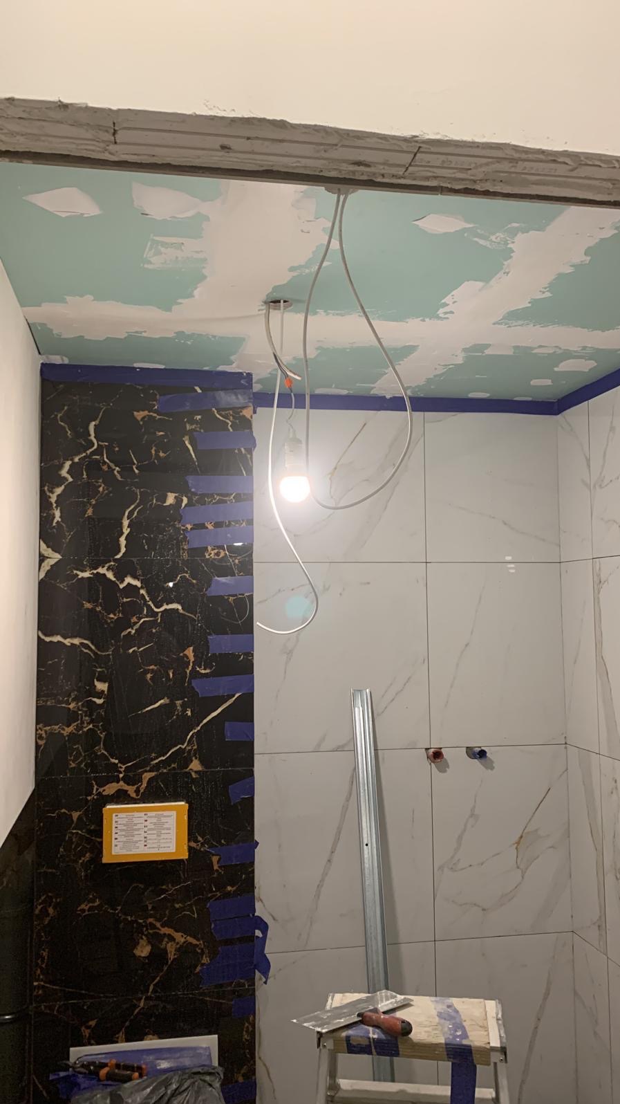 Łazienka w trakcie prac: częściowo ułożone płytki imitujące marmur w kolorze czarnym i białym, zabezpieczone niebieską taśmą malarską, widoczne narzędzia glazurnicze i aluminiowy profil, surowy...