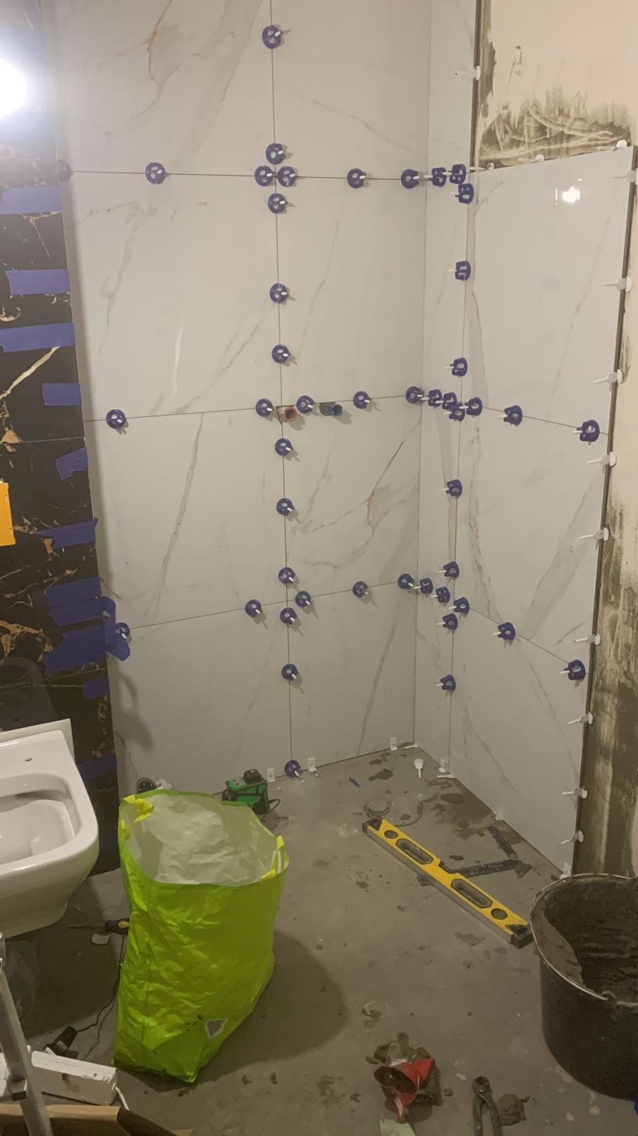 Montaż płytek imitujących marmur w rogu łazienki, z użyciem systemu poziomowania płytek, widoczny poziomica, wiadro z zaprawą i worek po kleju.