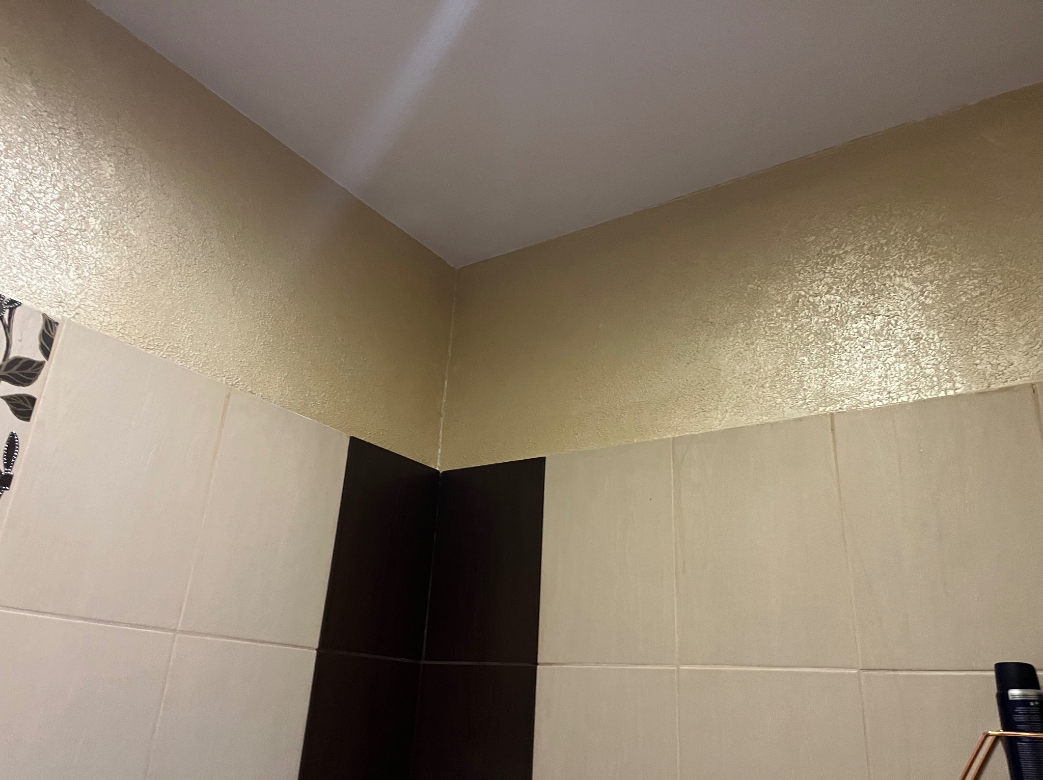 Fragment wykończonej łazienki: beżowe płytki, czarny pas płytek, dekoracyjna, złota struktura na ścianie pod sufitem, biały sufit
