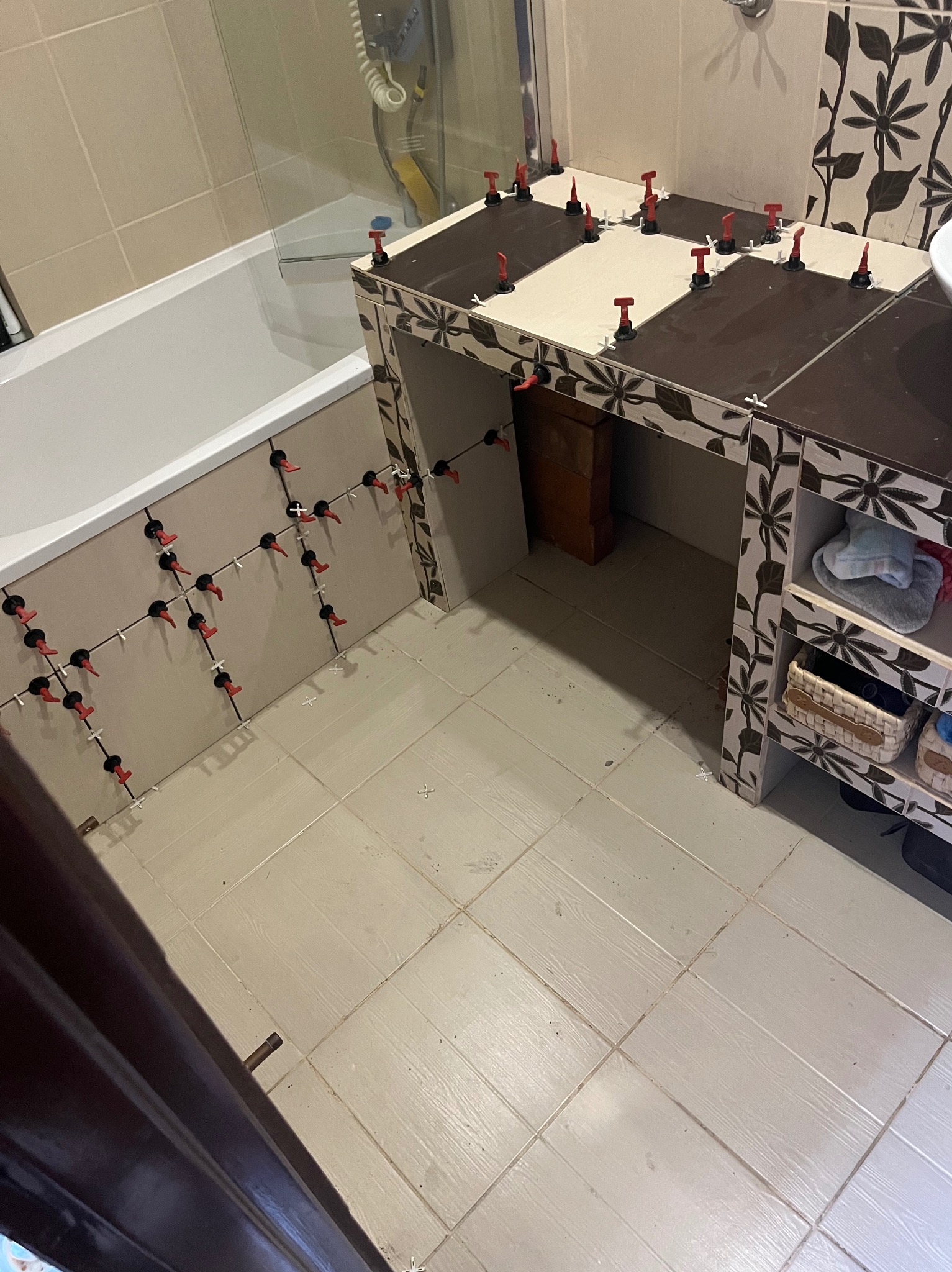 Łazienka w trakcie układania płytek: widoczne płytki przy wannie i na blacie pod umywalkę, z użyciem systemu poziomowania płytek (czerwone kliny i czarne podstawki).