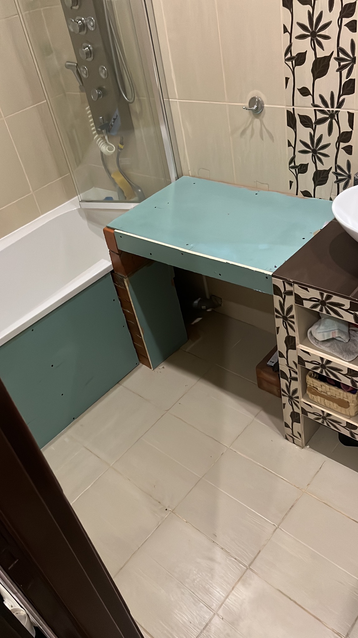 Niedokończona zabudowa w łazience: fragment wanny obudowany płytą gipsową, konstrukcja pod umywalkę z zielonym blatem, widoczne rury i płytki z motywem roślinnym.