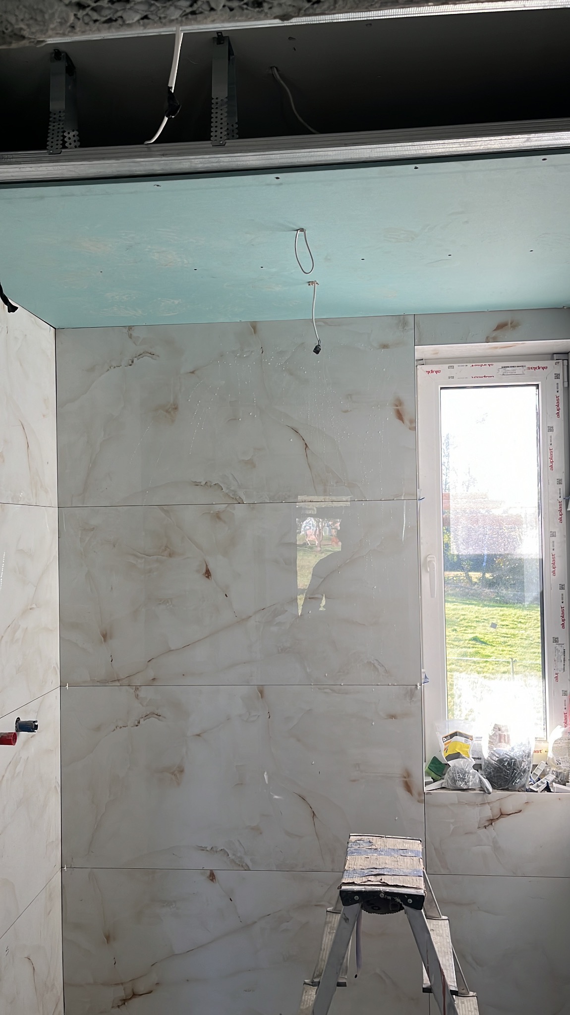 Ściana łazienki w trakcie układania płytek imitujących marmur, widoczna drabina malarska i okno z narzędziami, sufit z instalacją podwieszaną.