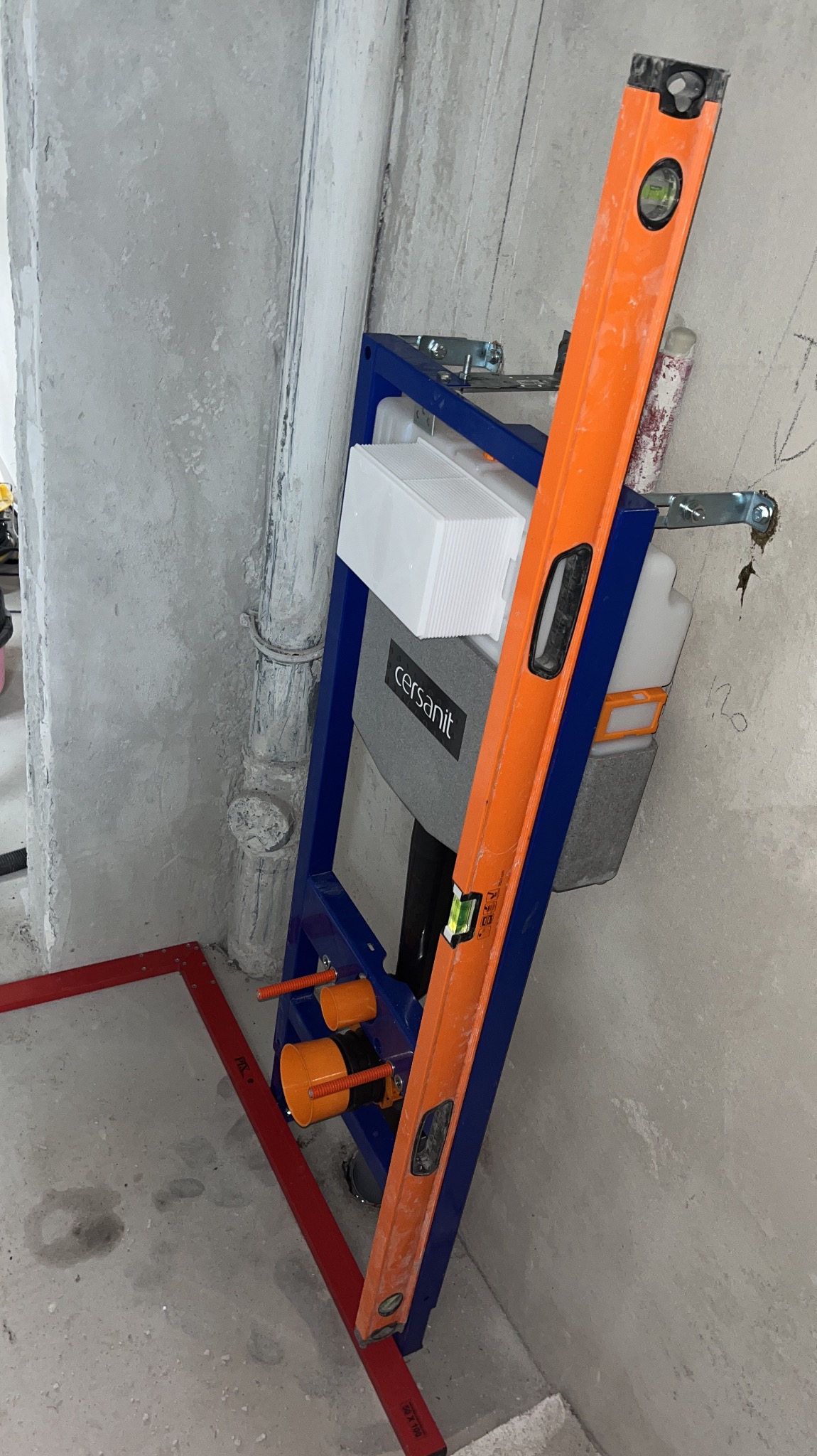 Stelaż podtynkowy Cersanit zamontowany przy ścianie, sprawdzany poziomicą, widoczne rury i fragment ramy z czerwonym obrysem na podłodze, surowy stan wykończenia.