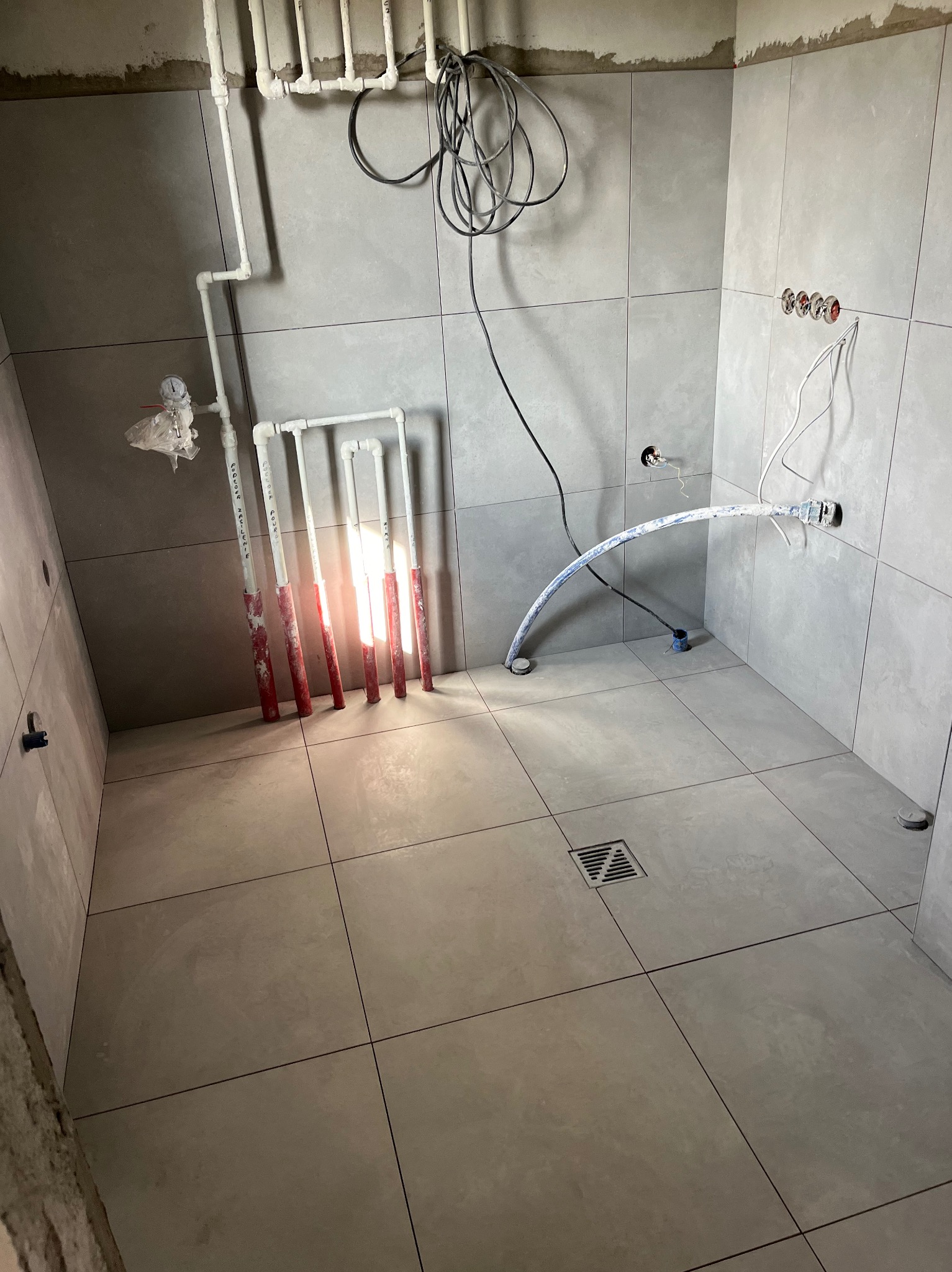 Szara łazienka w trakcie remontu, widoczne płytki na ścianach i podłodze, instalacja wodna w rurach z czerwonymi oznaczeniami, wystające kable elektryczne i odpływ liniowy w podłodze.