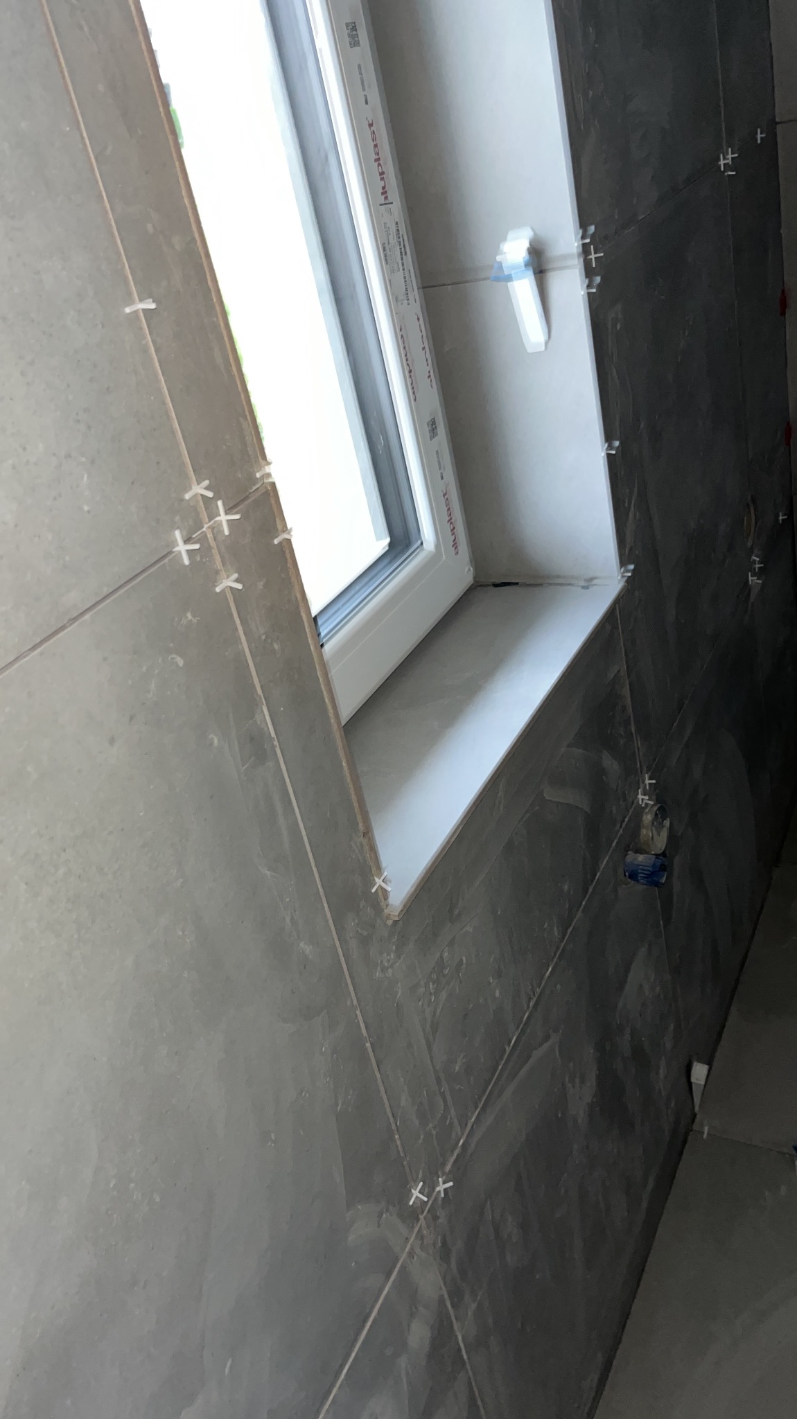 Układanie szarych płytek ceramicznych w łazience, widoczne krzyżyki dystansowe i okno z białym parapetem.