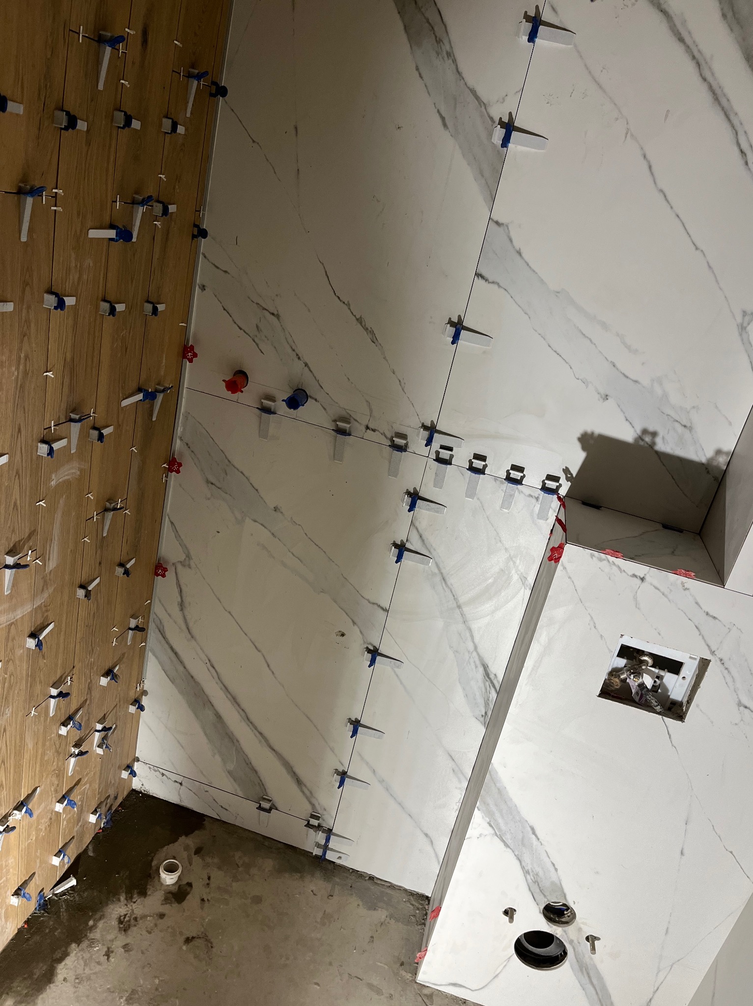 Świeżo ułożone, białe płytki imitujące marmur na ścianie, z systemem poziomowania płytek, widoczne również panele drewnopodobne na ścianie obok, w trakcie prac w Nowym Sączu.