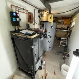 Nowoczesny piec grzewczy z czarną obudową i miedzianymi rurami w piwnicy, widoczne narzędzia i elementy instalacji, w tle zbiornik akumulacyjny i warsztat.