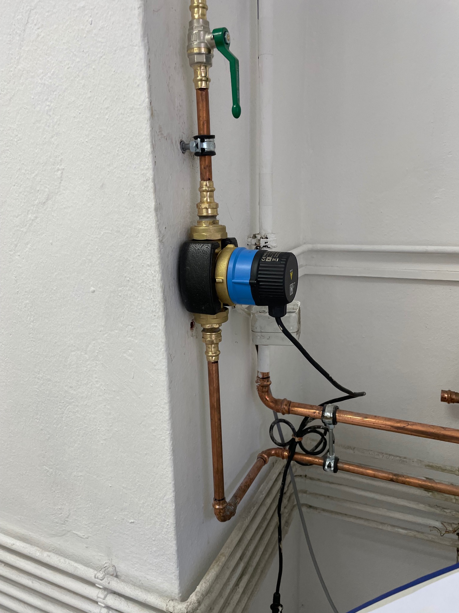 Zamontowana pompa obiegowa z niebieskim elementem na rurach miedzianych, zawór kulowy z zieloną rączką u góry, widoczne obejmy mocujące rury do ściany.