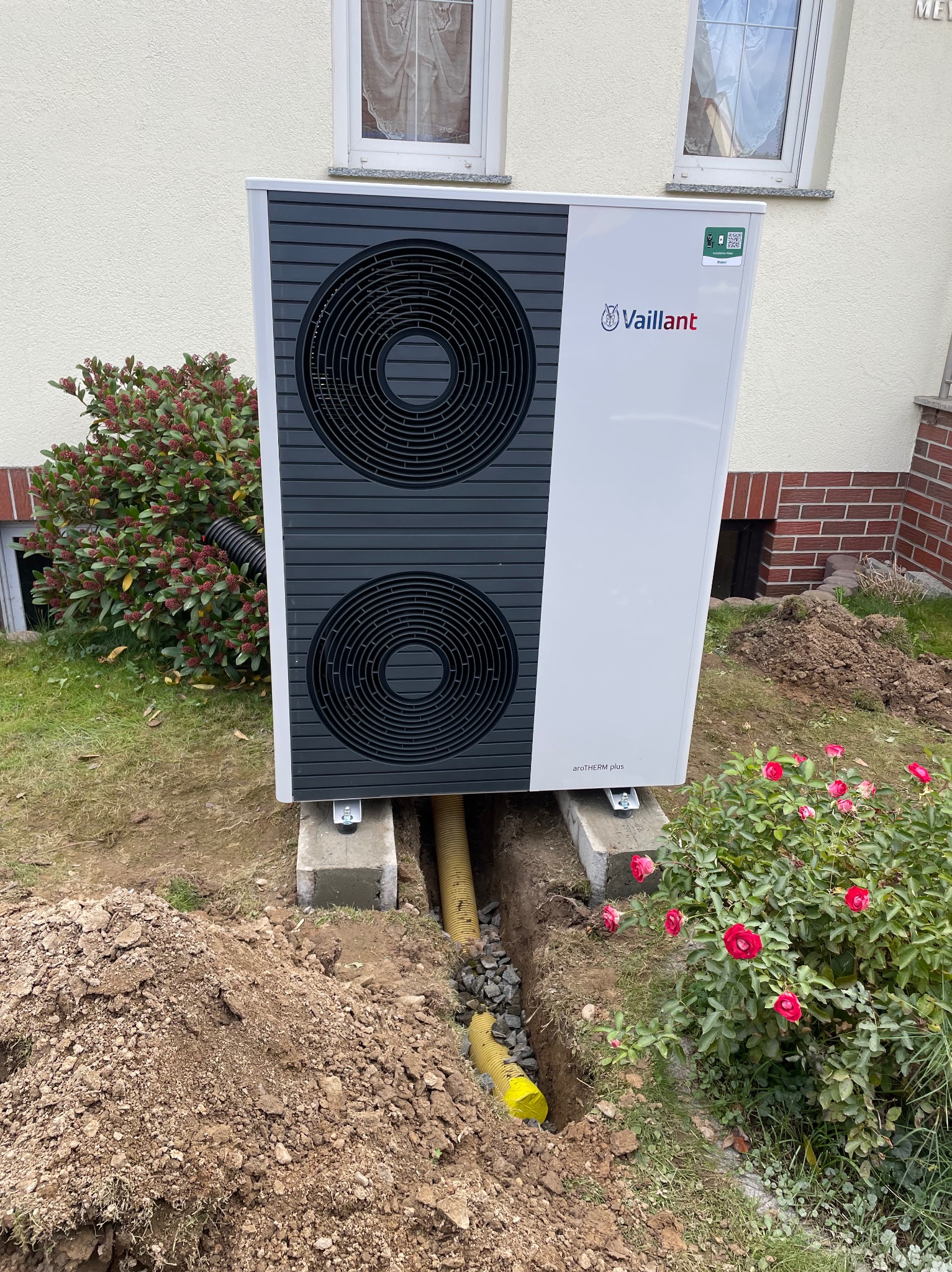 Biała pompa ciepła Vaillant AROTHERM plus, osadzona na betonowych wspornikach, z żółtą rurą odprowadzającą skropliny w ziemi, widoczna w ogrodzie przed domem.