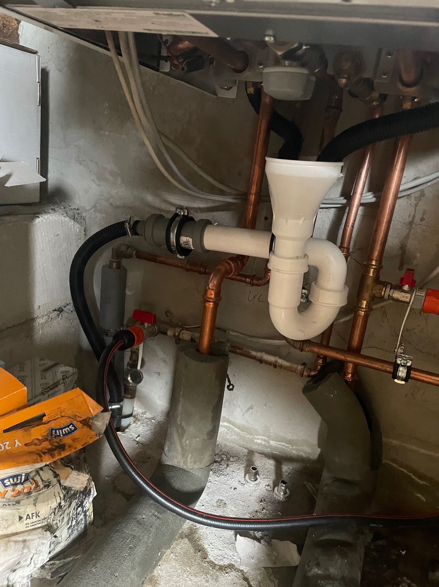 Fragment instalacji z miedzianych rur, białego syfonu i zaworów, widoczne elementy kotła i fragment opakowania materiałów budowlanych w surowym otoczeniu.