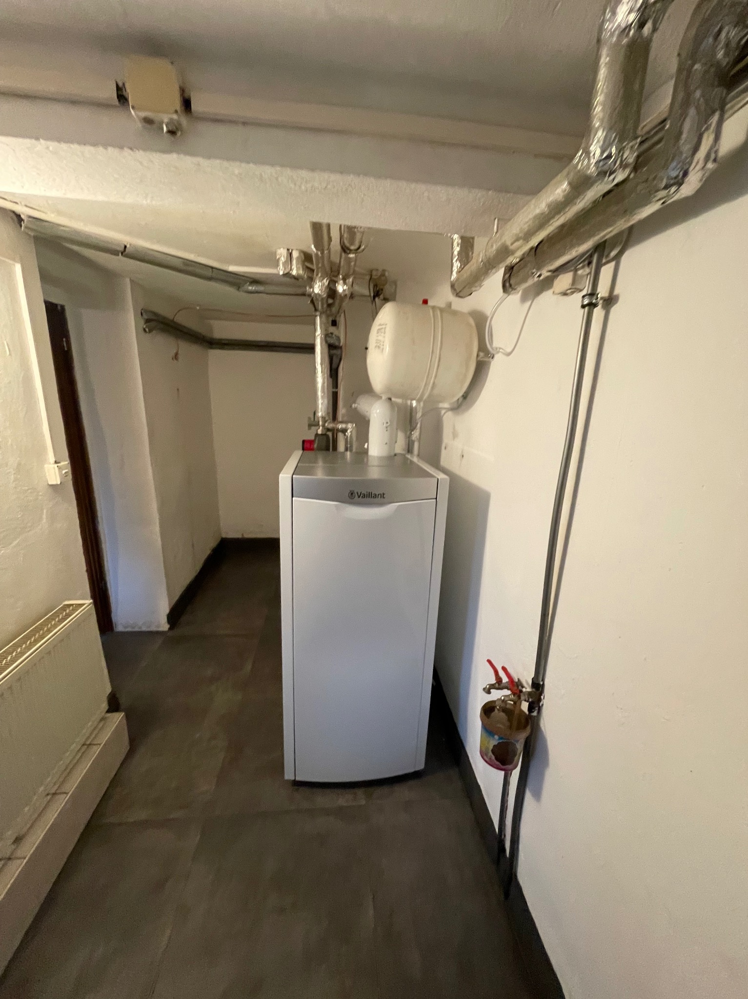 Nowoczesny, biały piec grzewczy Vaillant z białym zbiornikiem wyrównawczym w piwnicy z widocznymi rurami instalacji grzewczej, zabezpieczonymi folią aluminiową, oraz zawieszoną puszką z kranikami.
