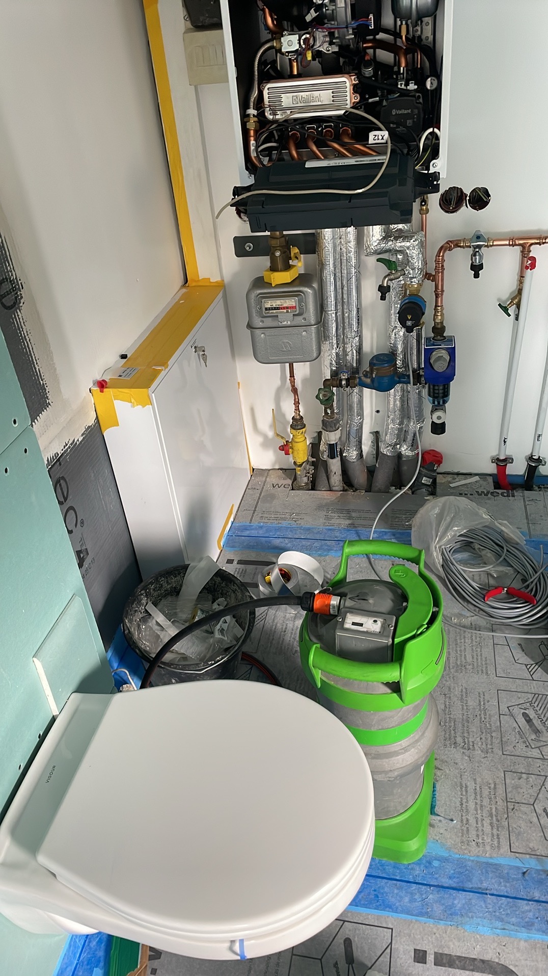 Wnętrze kotłowni w trakcie prac: widoczny wiszący piec gazowy Vaillant, rury instalacji grzewczej, licznik gazu, zielona maszyna do udrażniania rur oraz muszla klozetowa w folii ochronnej, co...