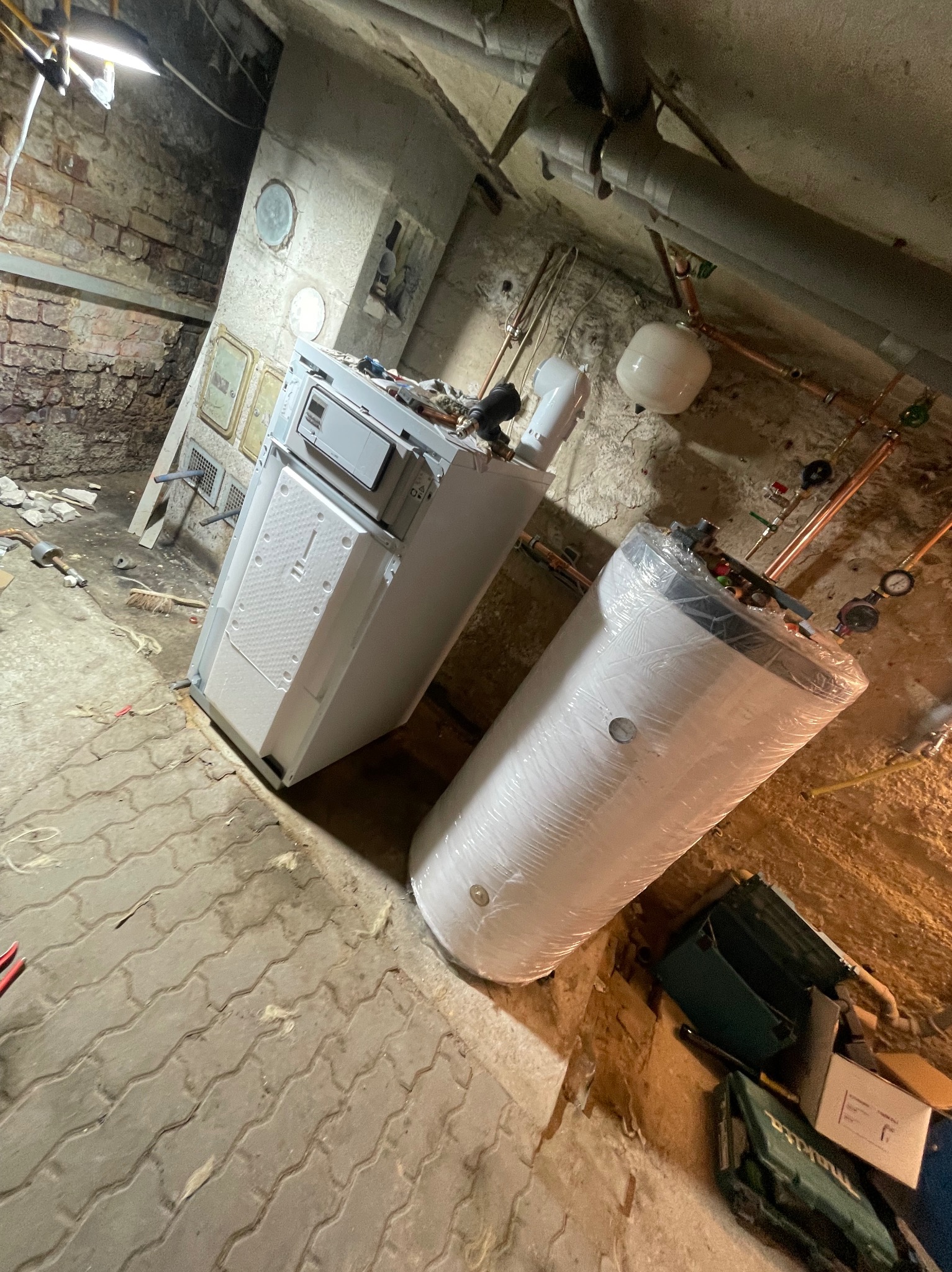 Nowy, biały piec grzewczy i zasobnik ciepłej wody w folii, czekające na montaż w piwnicy z ceglanymi ścianami i betonową podłogą.