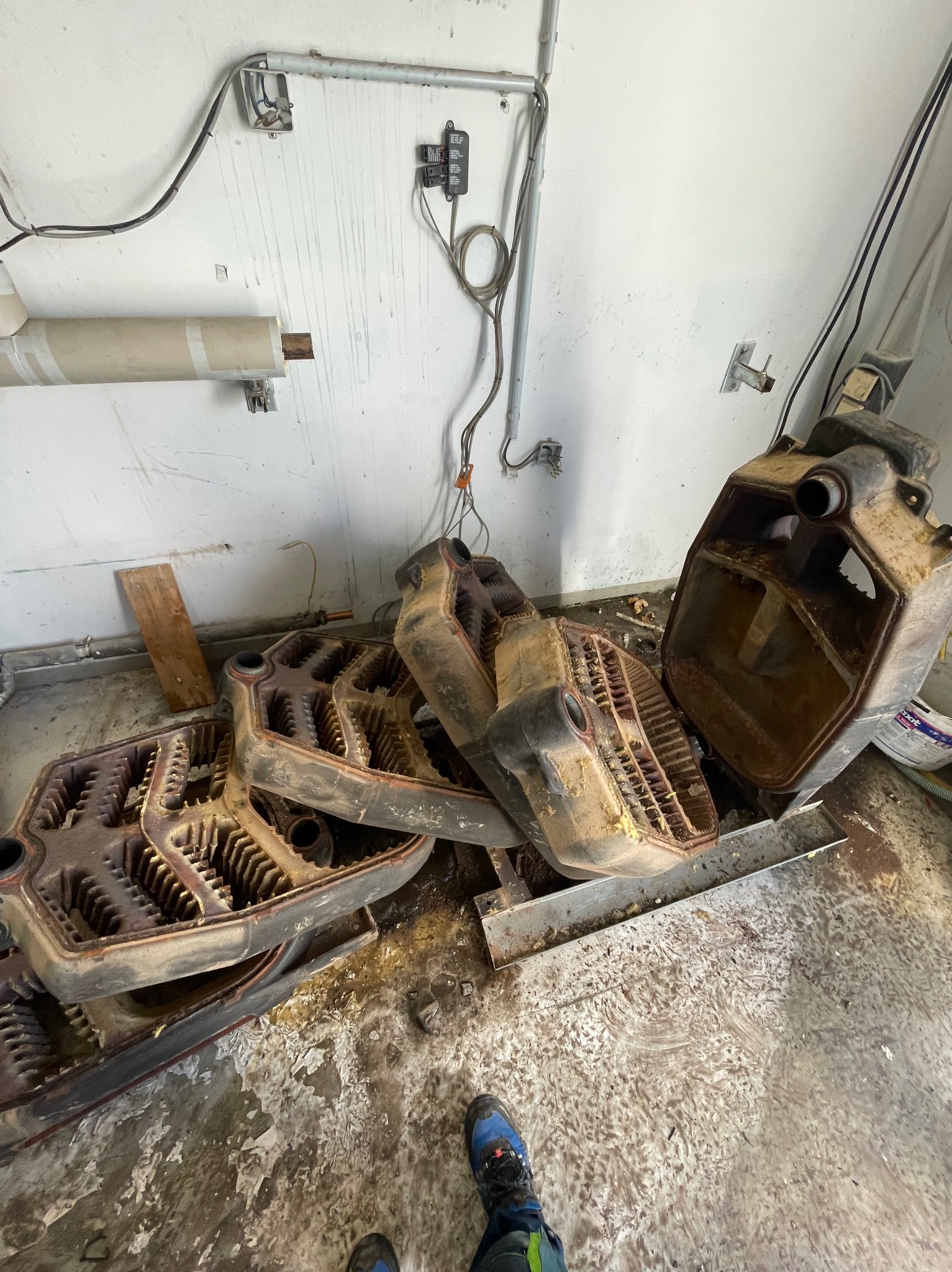 Zdemontowane, żeliwne segmenty grzejnika leżące na podłodze w pomieszczeniu z instalacjami elektrycznymi na ścianie. Widoczne ślady korozji i zabrudzeń.