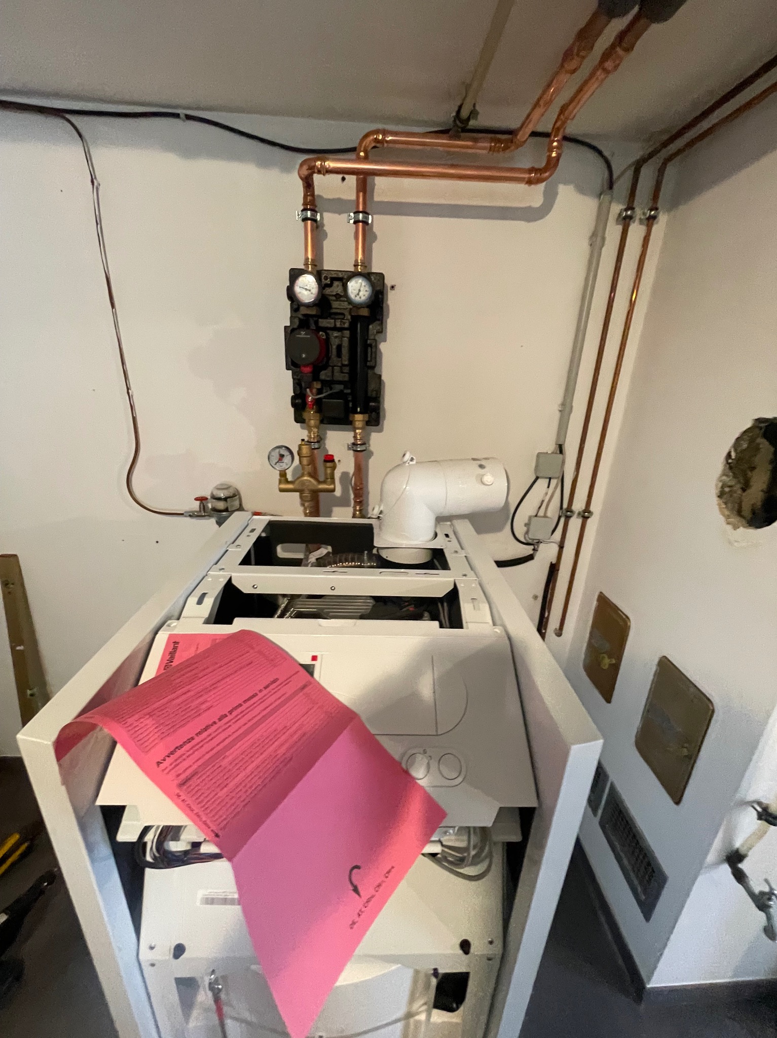 Otwarty piec grzewczy marki Vaillant z widocznymi elementami instalacji miedzianej na ścianie, instrukcja w kolorze różowym częściowo zasłaniająca wnętrze pieca.