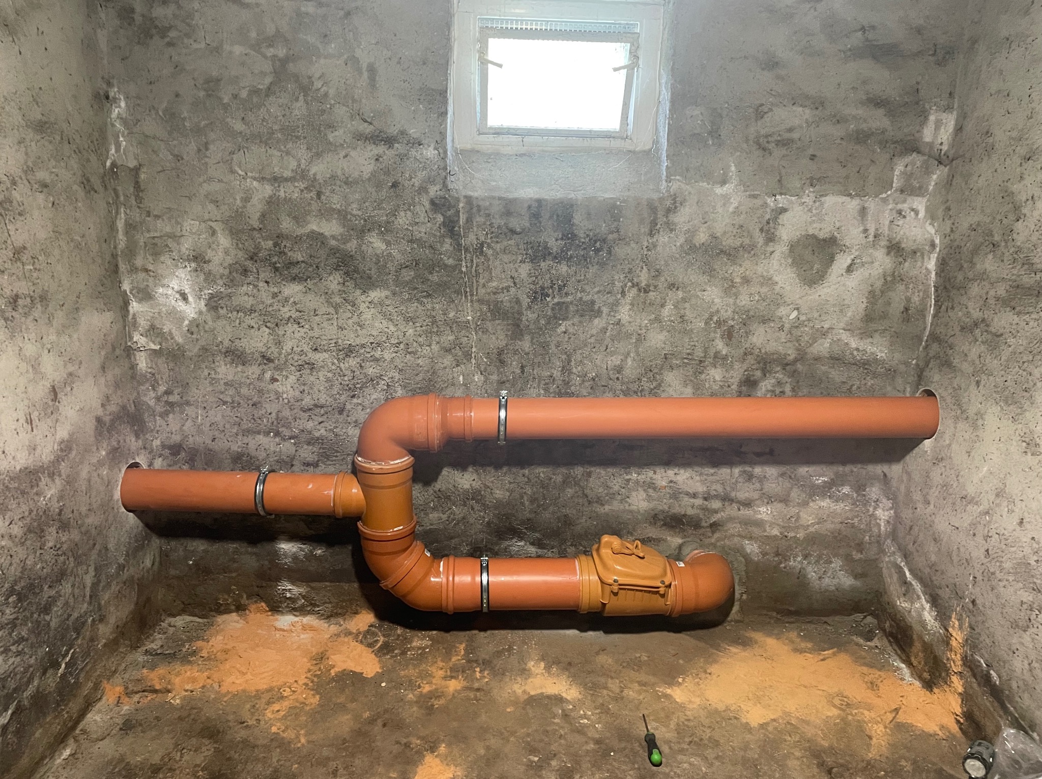 Pomarańczowe rury kanalizacyjne z zaworem zwrotnym zamontowane w betonowej piwnicy z małym oknem, widoczny ślad po wierceniu w ścianie i śrubokręt na podłodze.