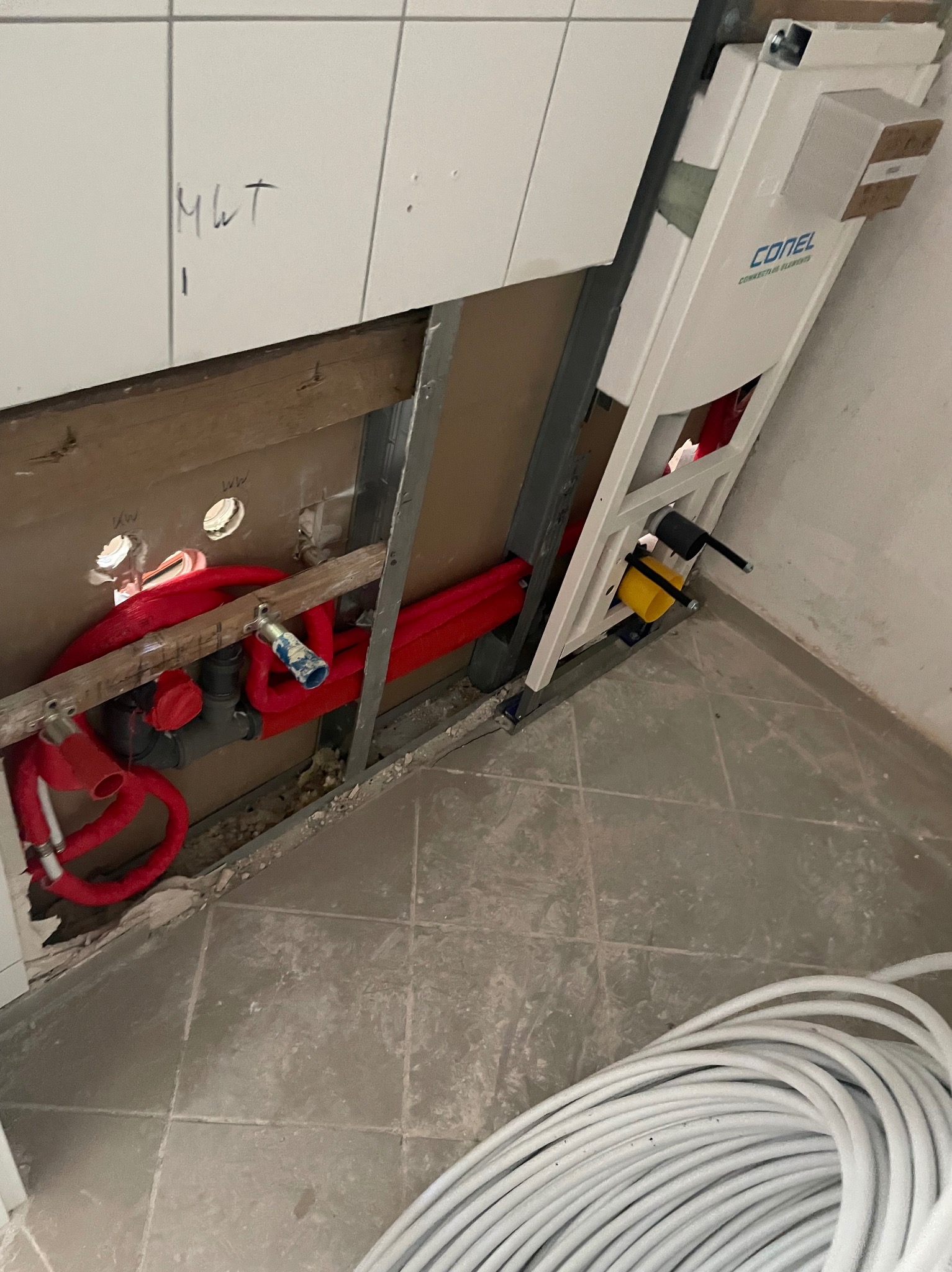 Częściowo odsłonięta instalacja podtynkowa WC z czerwonymi rurami doprowadzającymi wodę, widoczny stelaż marki Conel, białe płytki z napisem 'MWT' i zwój białych rur na podłodze.