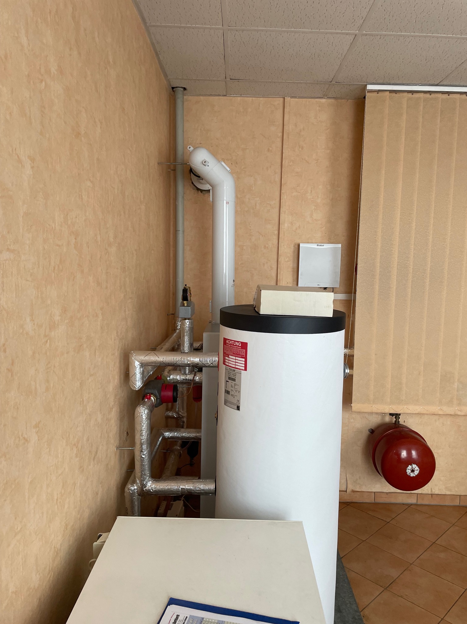 Biały zasobnik ciepłej wody z rurami w izolacji, podłączony do instalacji grzewczej w pomieszczeniu z beżowymi ścianami i brązową podłogą.