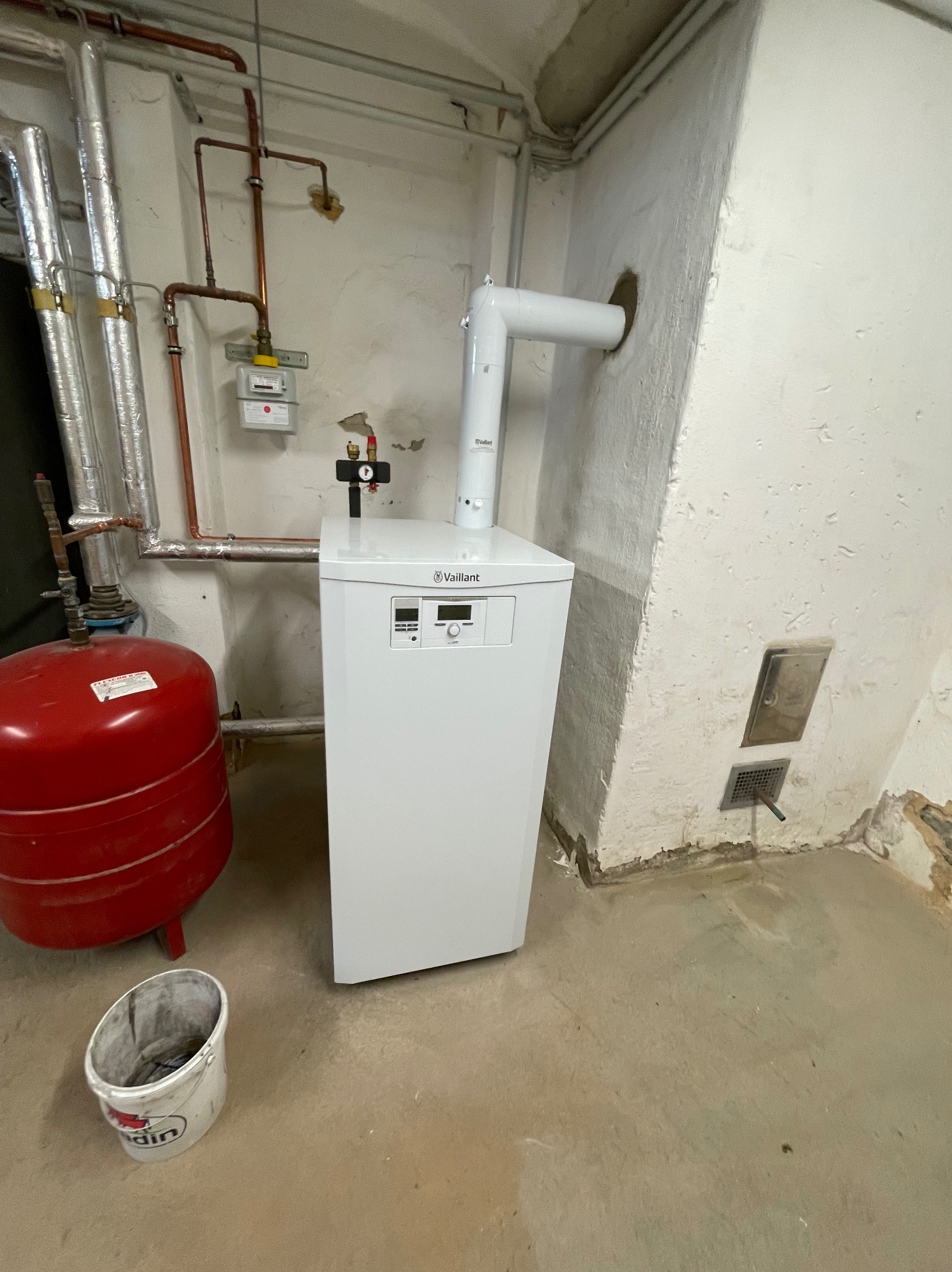 Nowy, biały piec gazowy marki Vaillant, z podłączonym kominem i instalacją hydrauliczną, zamontowany w pomieszczeniu z widocznymi śladami wilgoci na ścianach i podłodze, obok czerwony zbiornik...