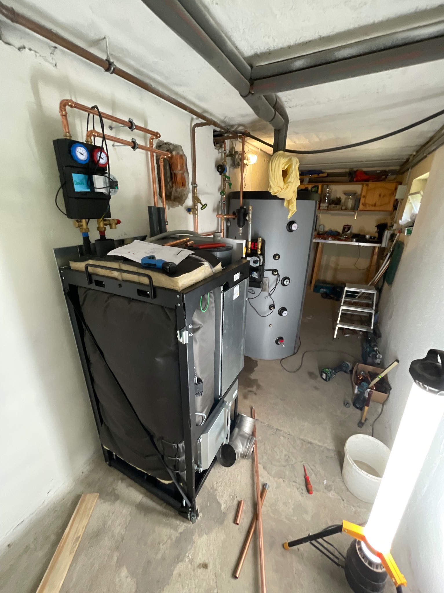 Nowoczesny piec grzewczy z czarną obudową i miedzianymi rurami w piwnicy, widoczne narzędzia i elementy instalacji, w tle zbiornik akumulacyjny i warsztat.