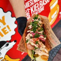 Hot dog z kiełbasą, sosem, czerwoną cebulą i zielonym szczypiorkiem, podawany w papierowej foremce, trzymany w ręce w czarnej rękawiczce na tle czerwonego food trucka z grafiką.