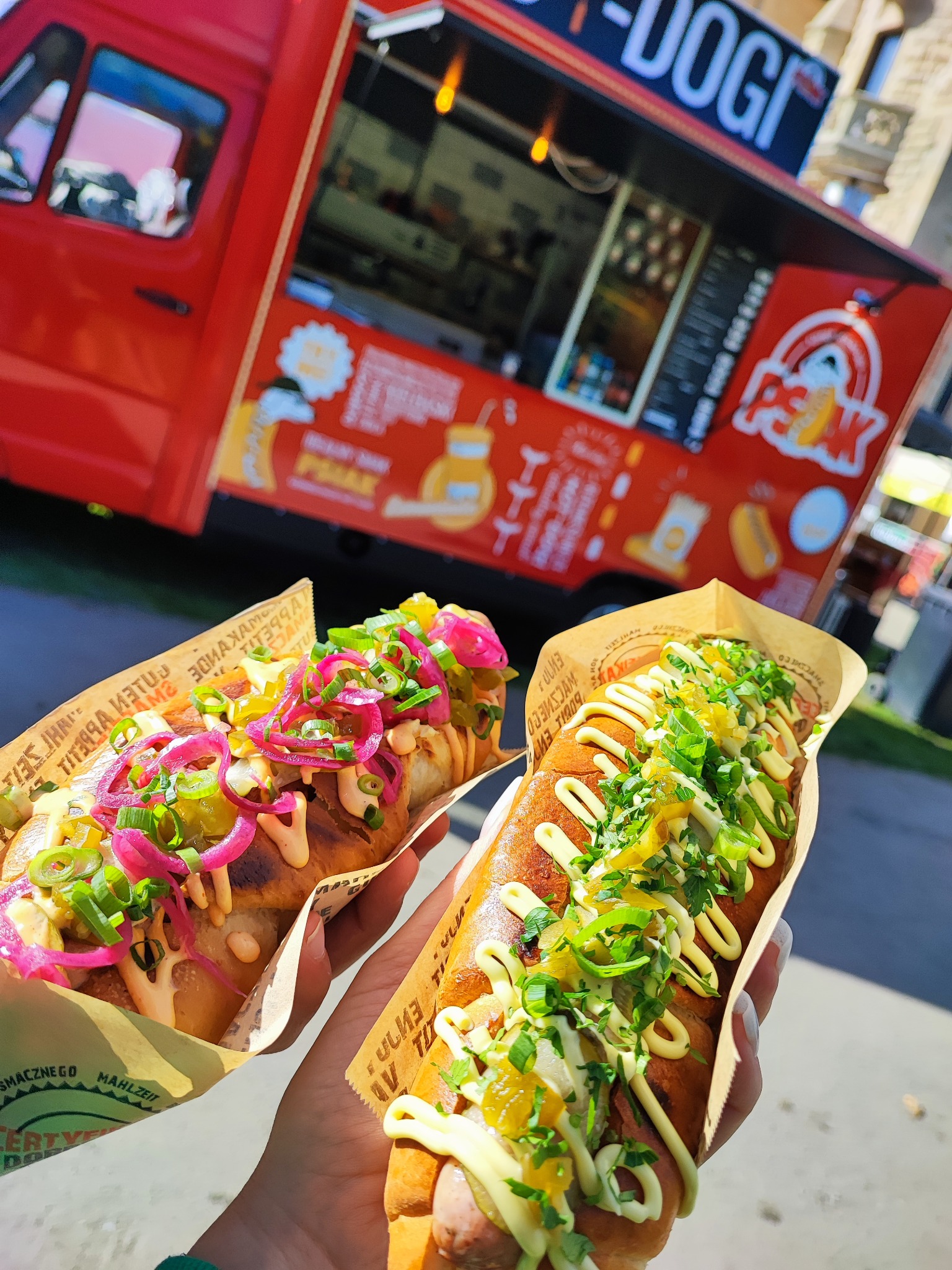 Dwa hot dogi z różnymi dodatkami (czerwona cebula i zielone zioła) trzymane w rękach na tle czerwonego food trucka z logo 'Peak'