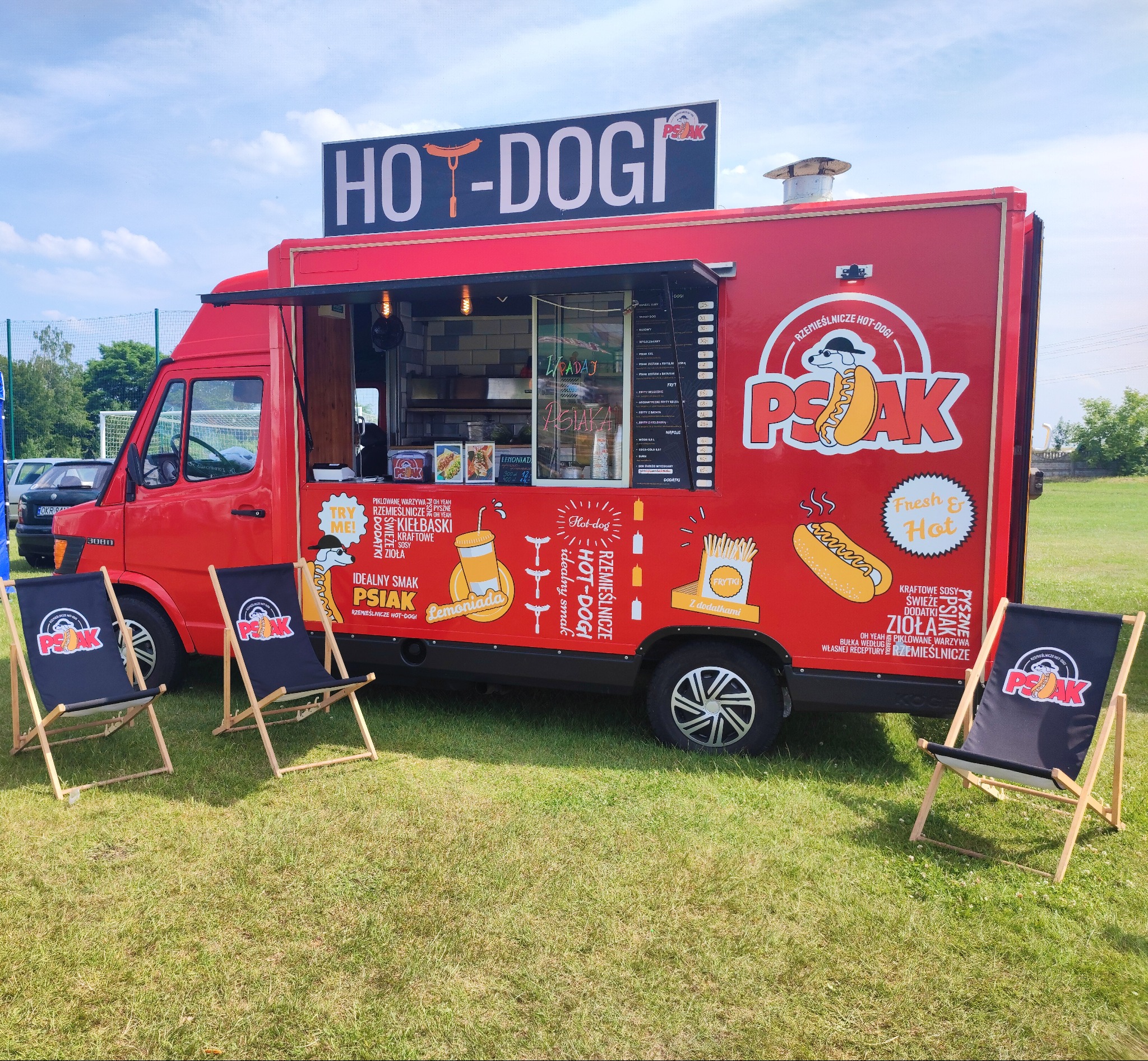 Czerwony food truck z szyldem 'Hot-Dogi' na zielonej trawie, z trzema leżakami z logo firmy 'Psak' przed nim, sugerującymi miejsce do odpoczynku dla klientów.