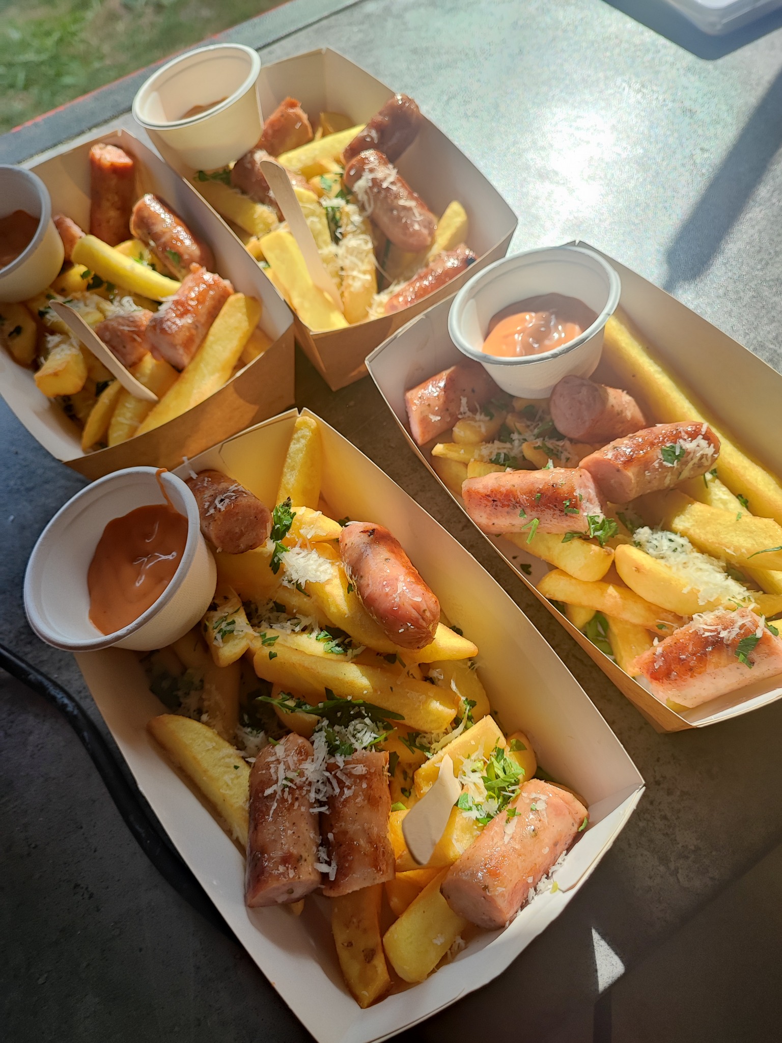 Cztery porcje frytek z kawałkami kiełbasy, posypane serem i ziołami, podane w papierowych tackach z sosem w małych kubeczkach.