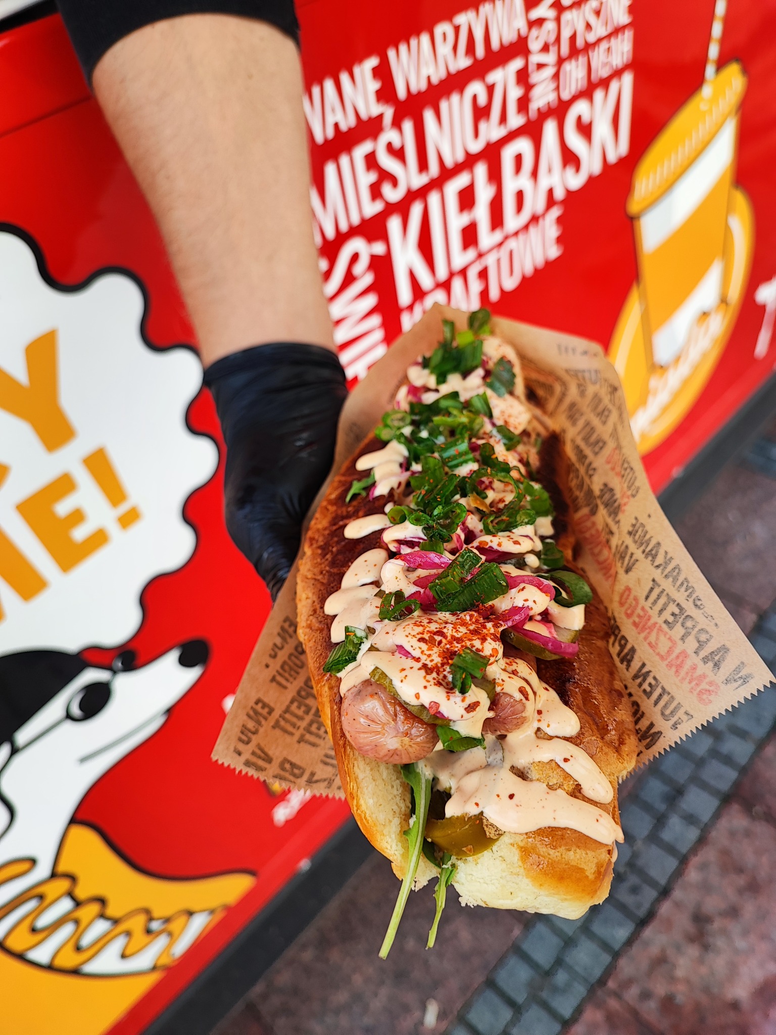 Hot dog z kiełbasą, sosem, czerwoną cebulą i zielonym szczypiorkiem, podawany w papierowej foremce, trzymany w ręce w czarnej rękawiczce na tle czerwonego food trucka z grafiką.