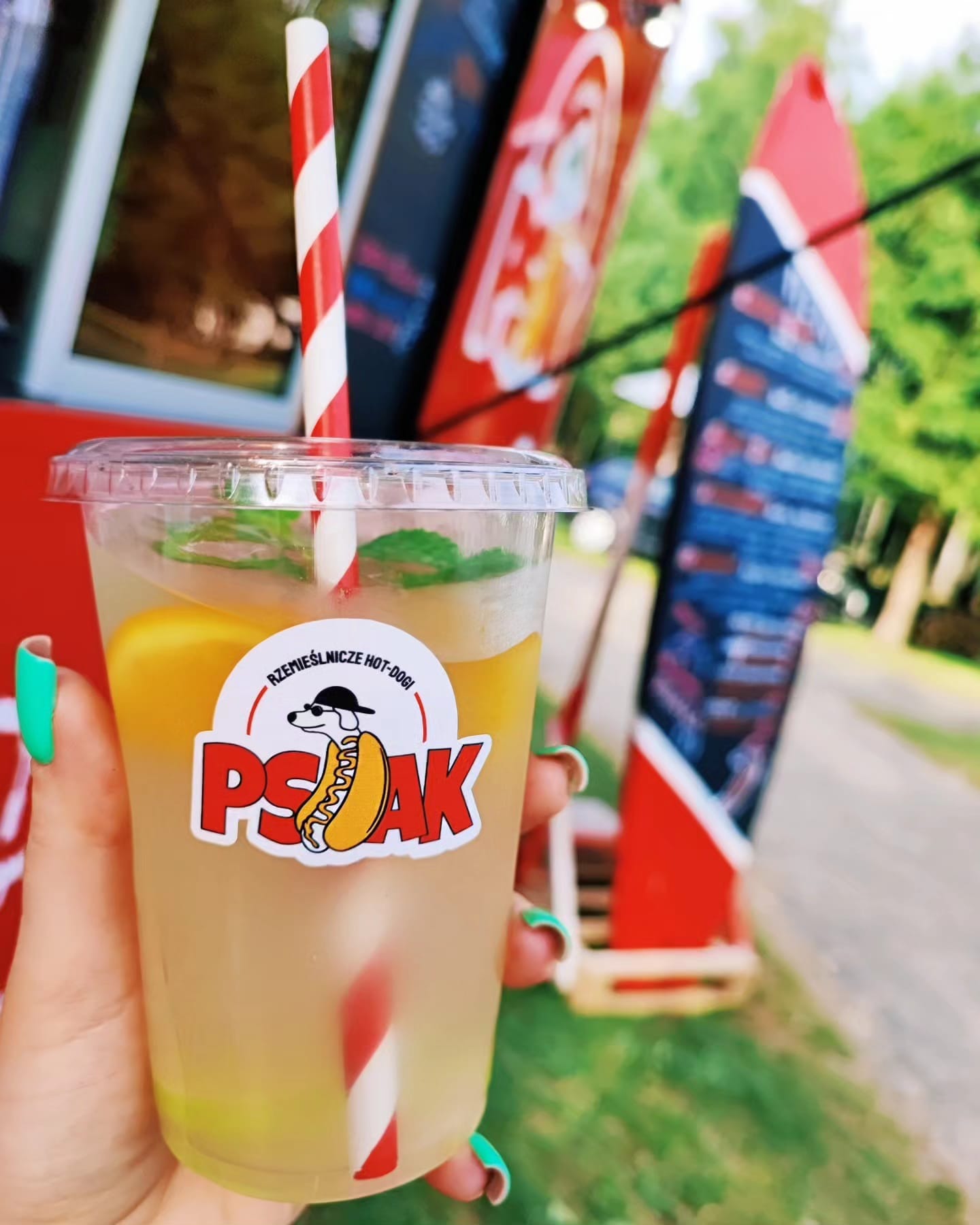 Ręka trzyma plastikowy kubek lemoniady z plasterkiem cytryny, miętą i słomką w biało-czerwone paski, na kubku naklejka z logo 'Psak - rzemieślnicze hot-dogi', w tle food truck.
