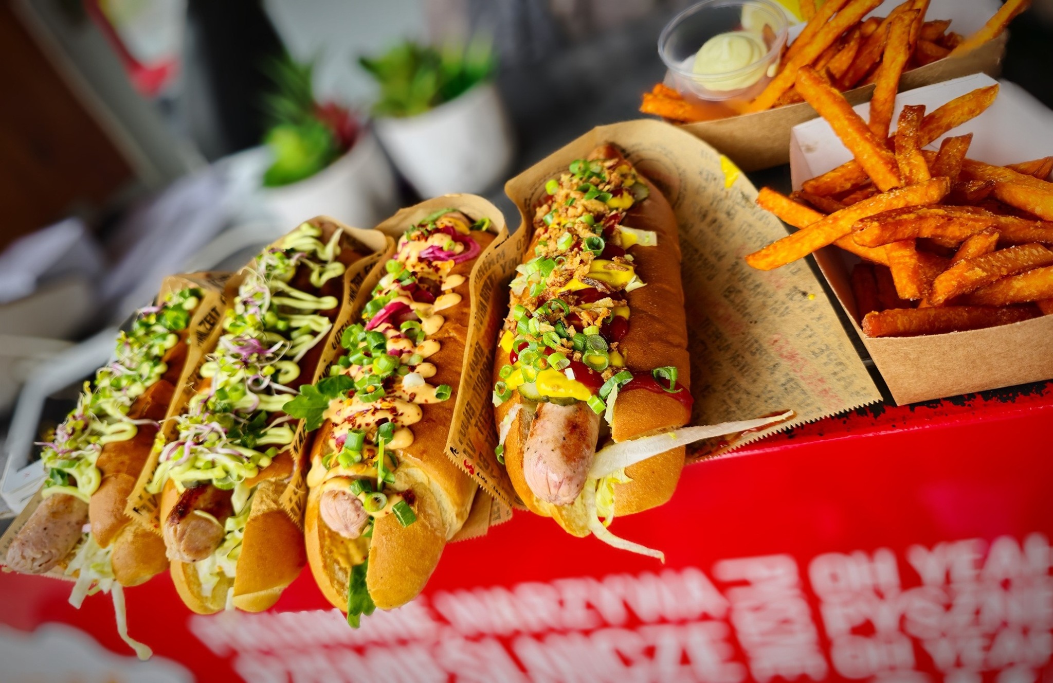 Cztery hot dogi z różnymi dodatkami (kiełki, sosy, prażona cebula, warzywa) i frytki w tekturowych pojemnikach, ułożone na czerwonej powierzchni z napisem.